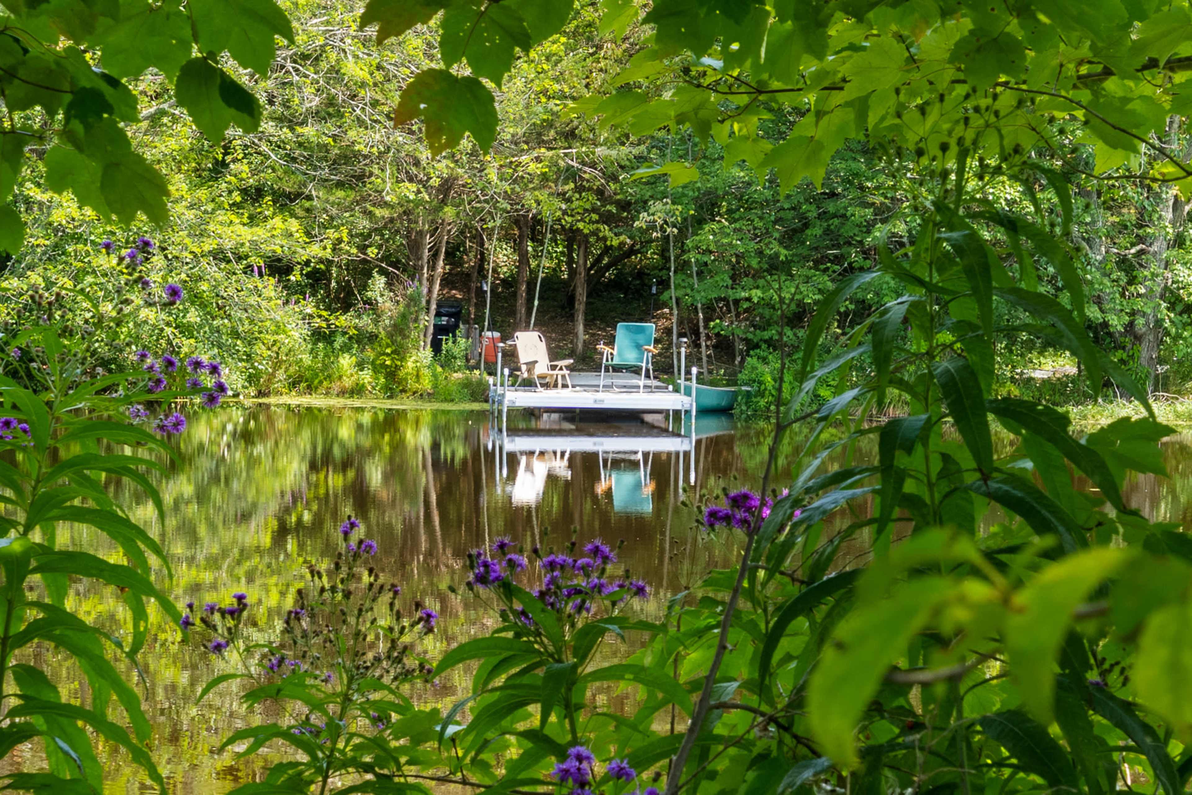 A small dock with two chairs is situated on a tranquil pond, framed by greenery and flowers.