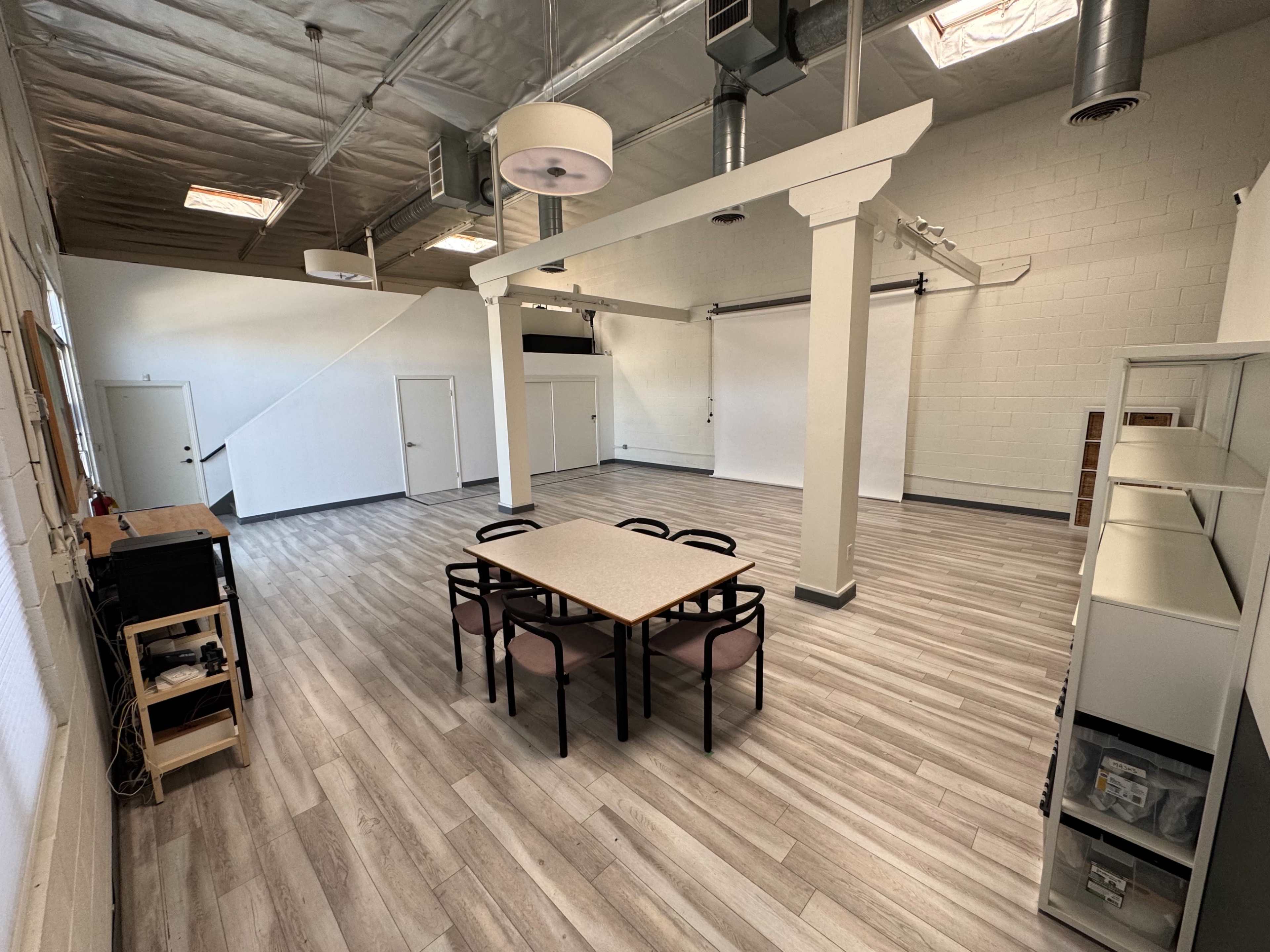 The image shows a spacious, well-lit room with a central table surrounded by chairs, white walls, and light wooden flooring.