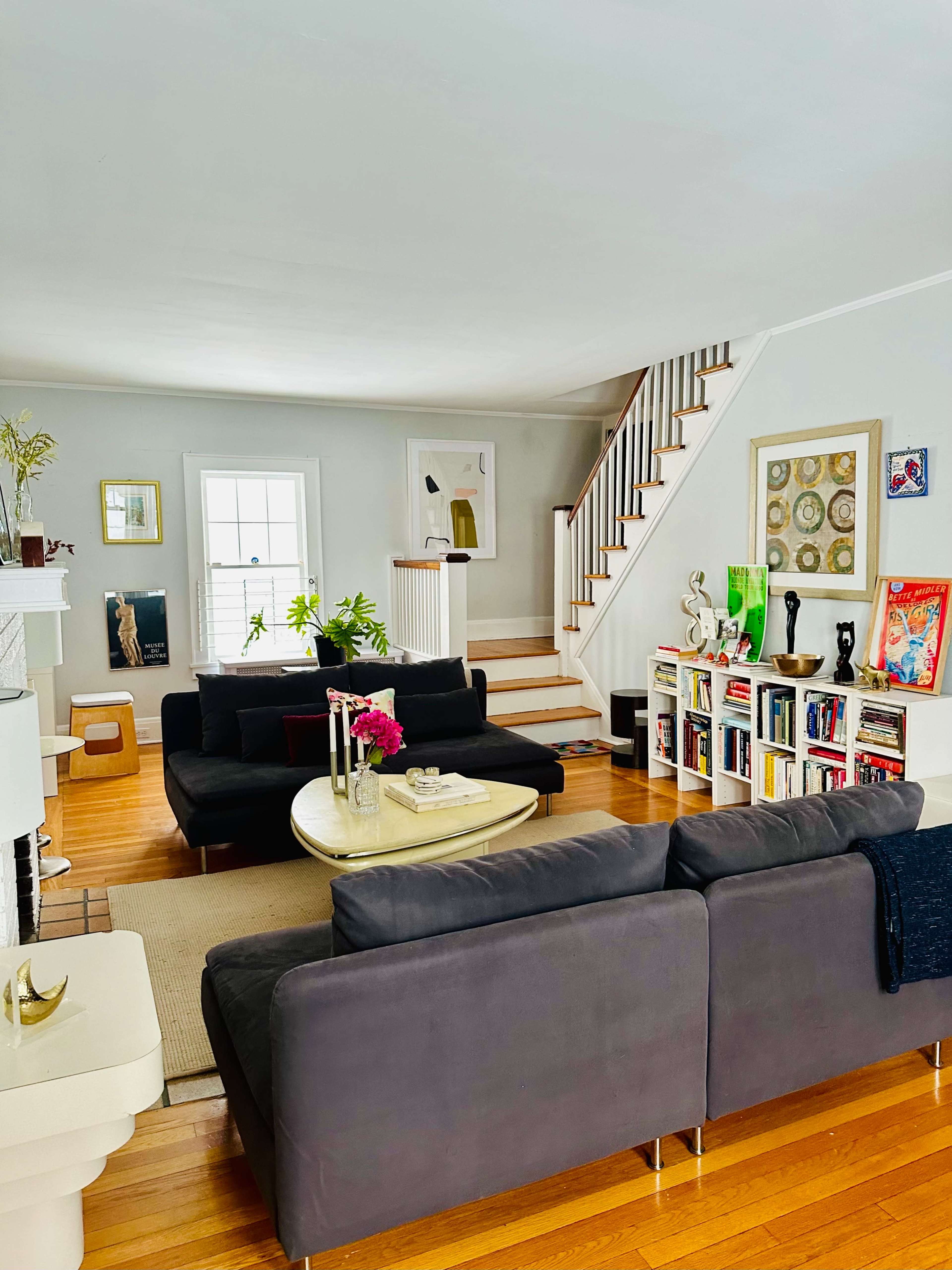 The living room features two dark sofas facing a central coffee table, with a staircase and a bookshelf in the background.