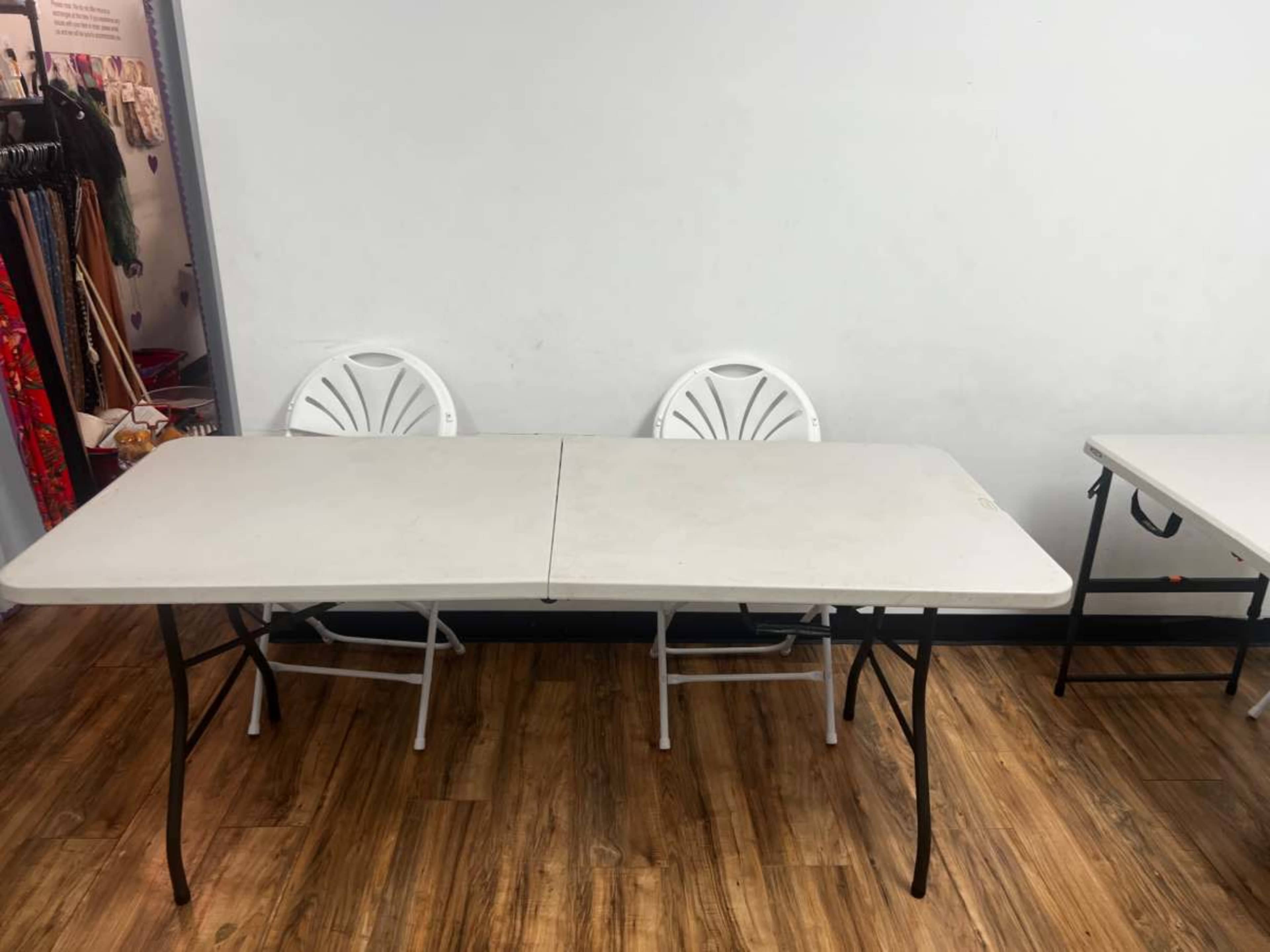 The image shows a rectangular folding table with two white chairs on one side, placed against a wall in a room with wooden flooring.