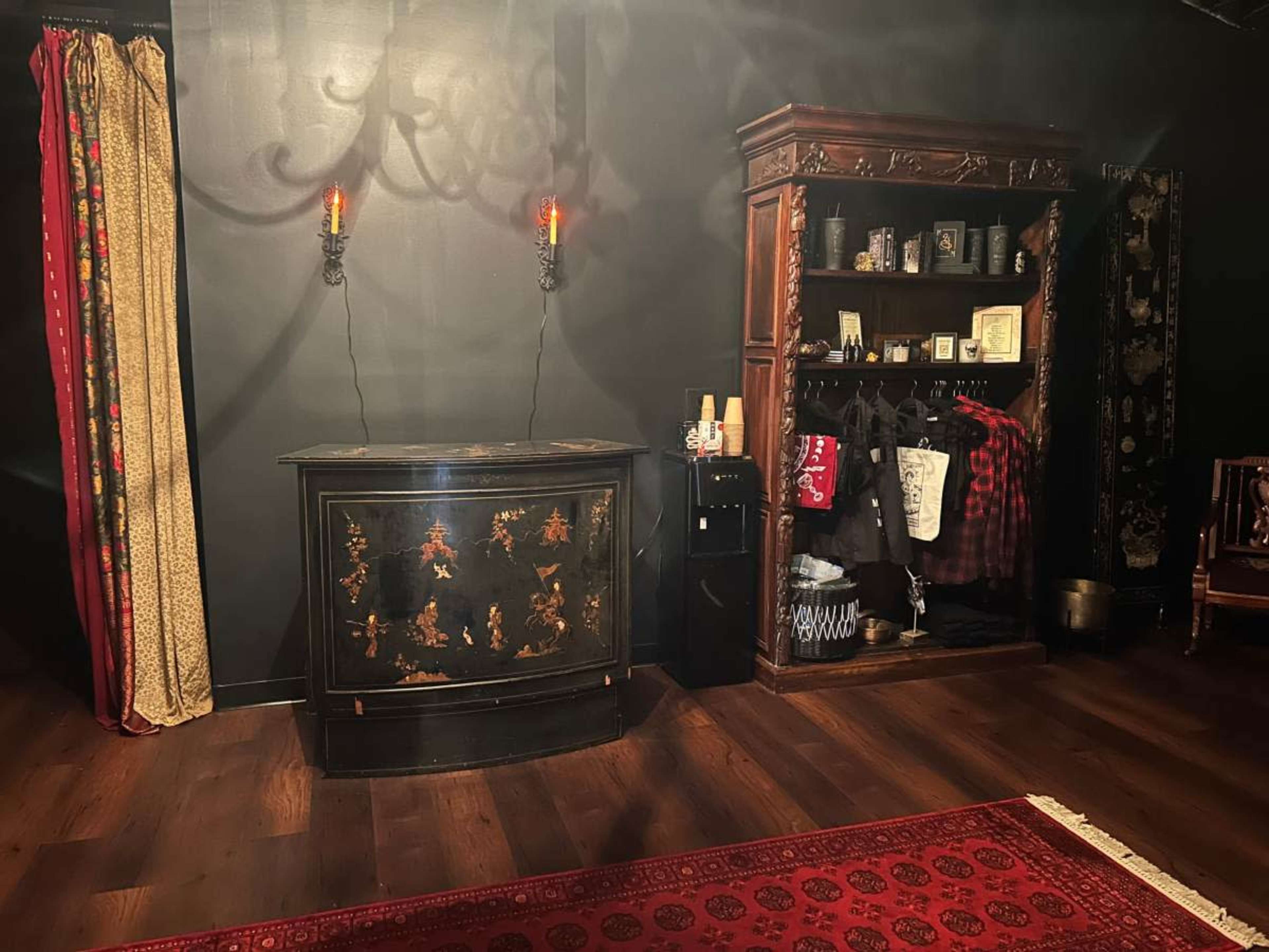 The image shows a dimly lit room featuring a dark wooden cabinet adorned with floral designs, a side table with red curtains, and a clothes rack displaying various garments against a shadowy wall.