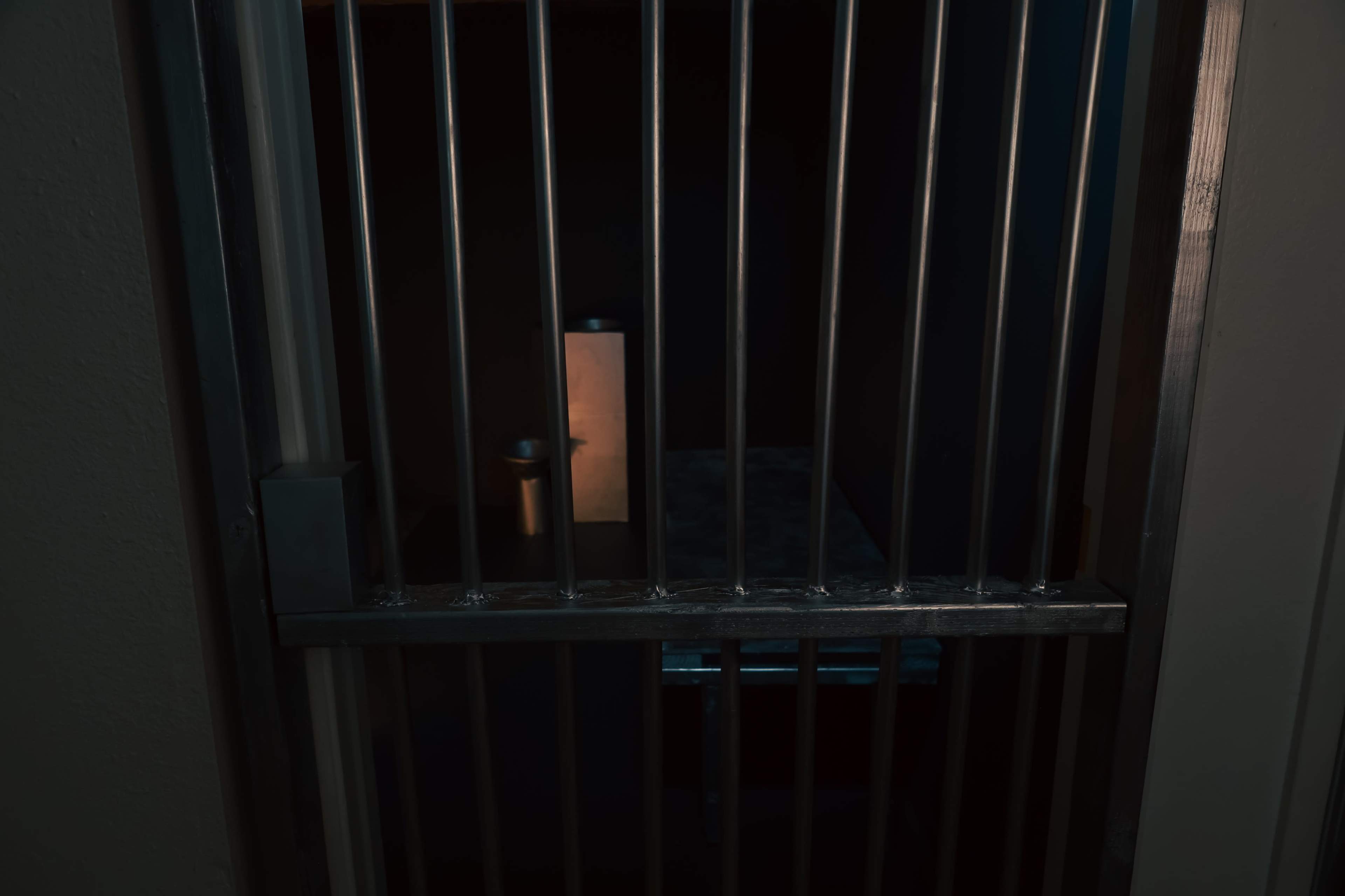 The image shows the bars of a prison cell with a dimly lit interior, featuring a small table and a cup.