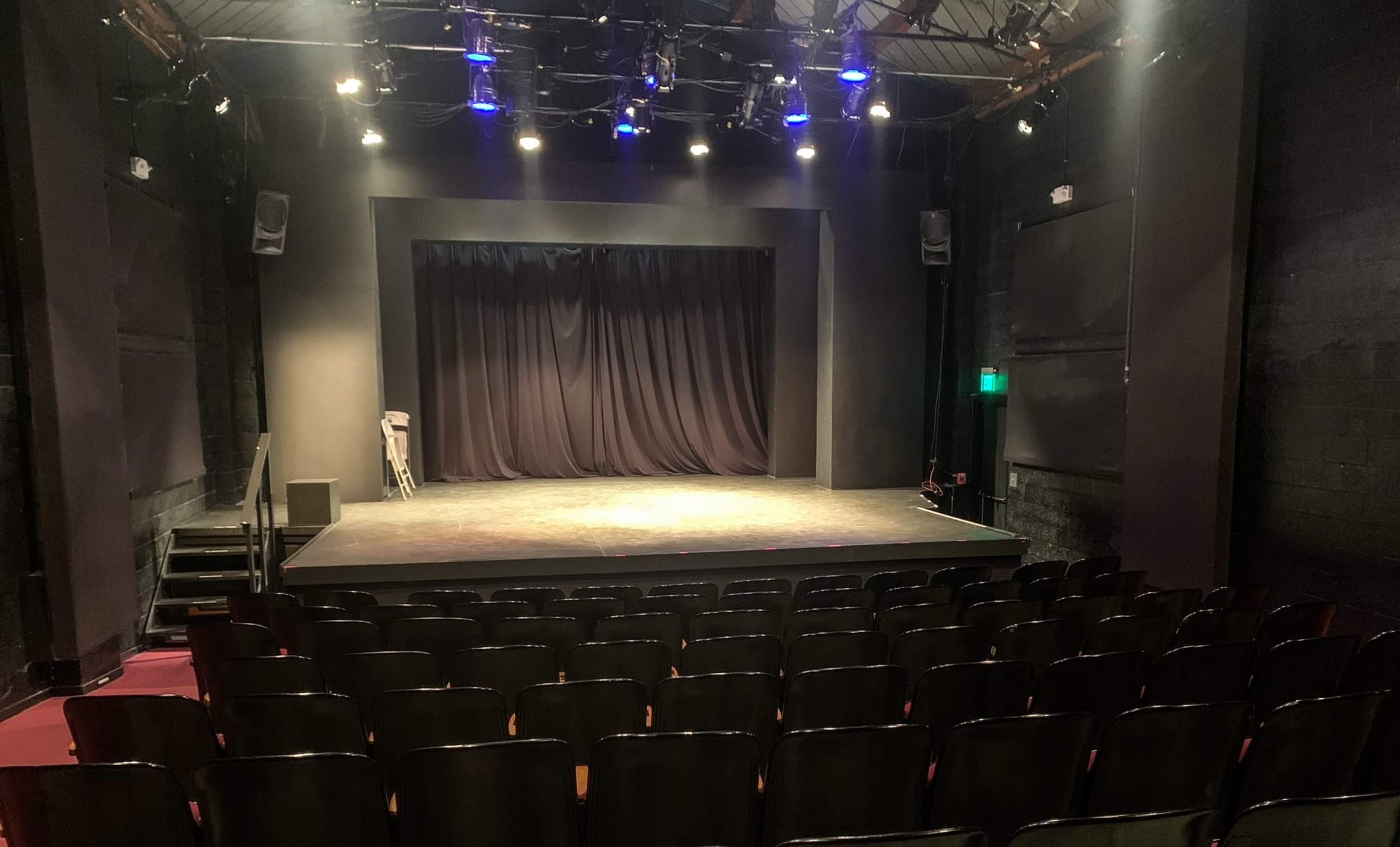 A darkened theater stage with a curtain in the background and rows of empty black seats in front.