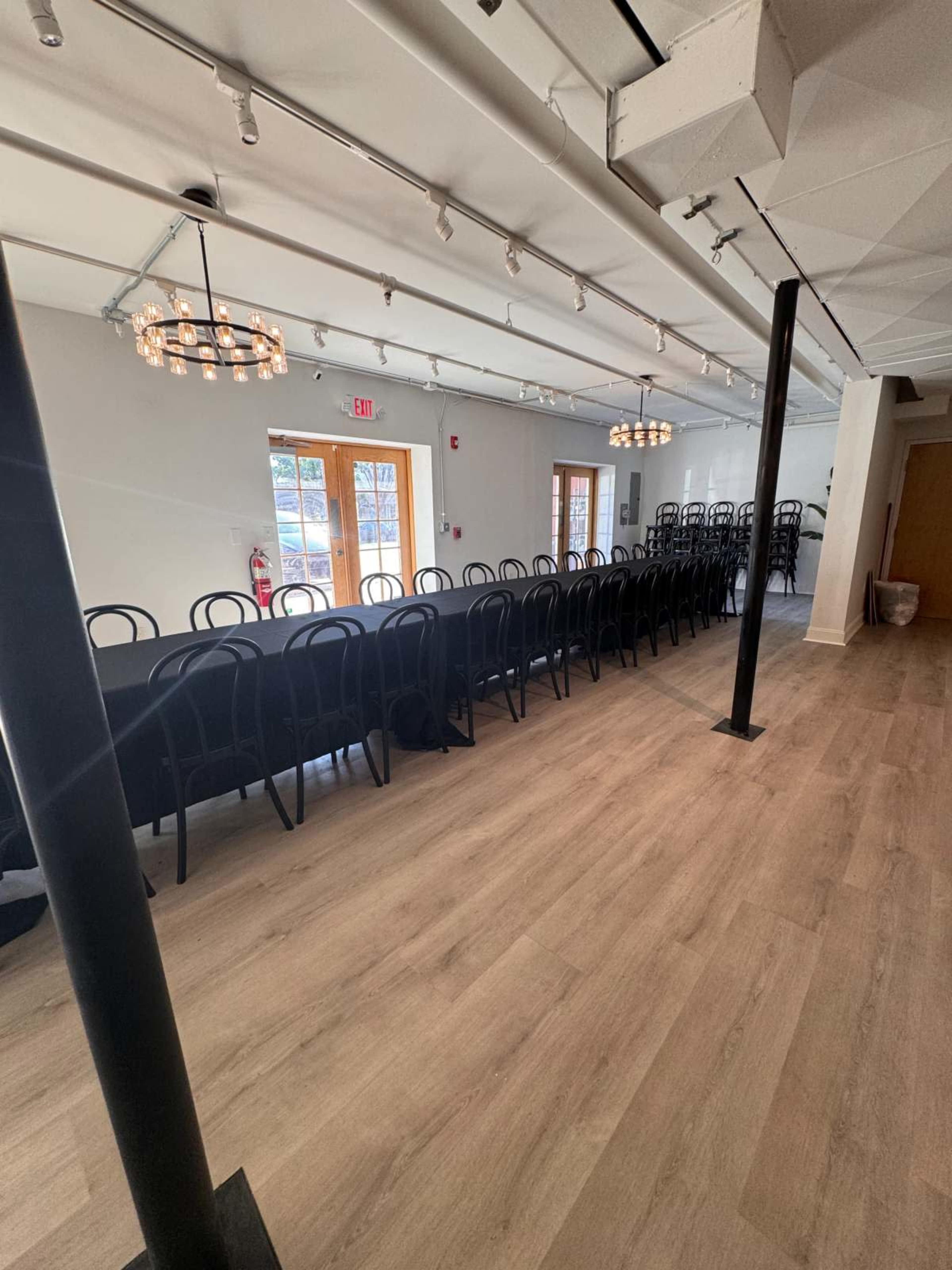 The image shows a well-lit indoor space set up with an elongated table covered in a black cloth and surrounded by black chairs, with large windows and two circular light fixtures hanging from the ceiling.