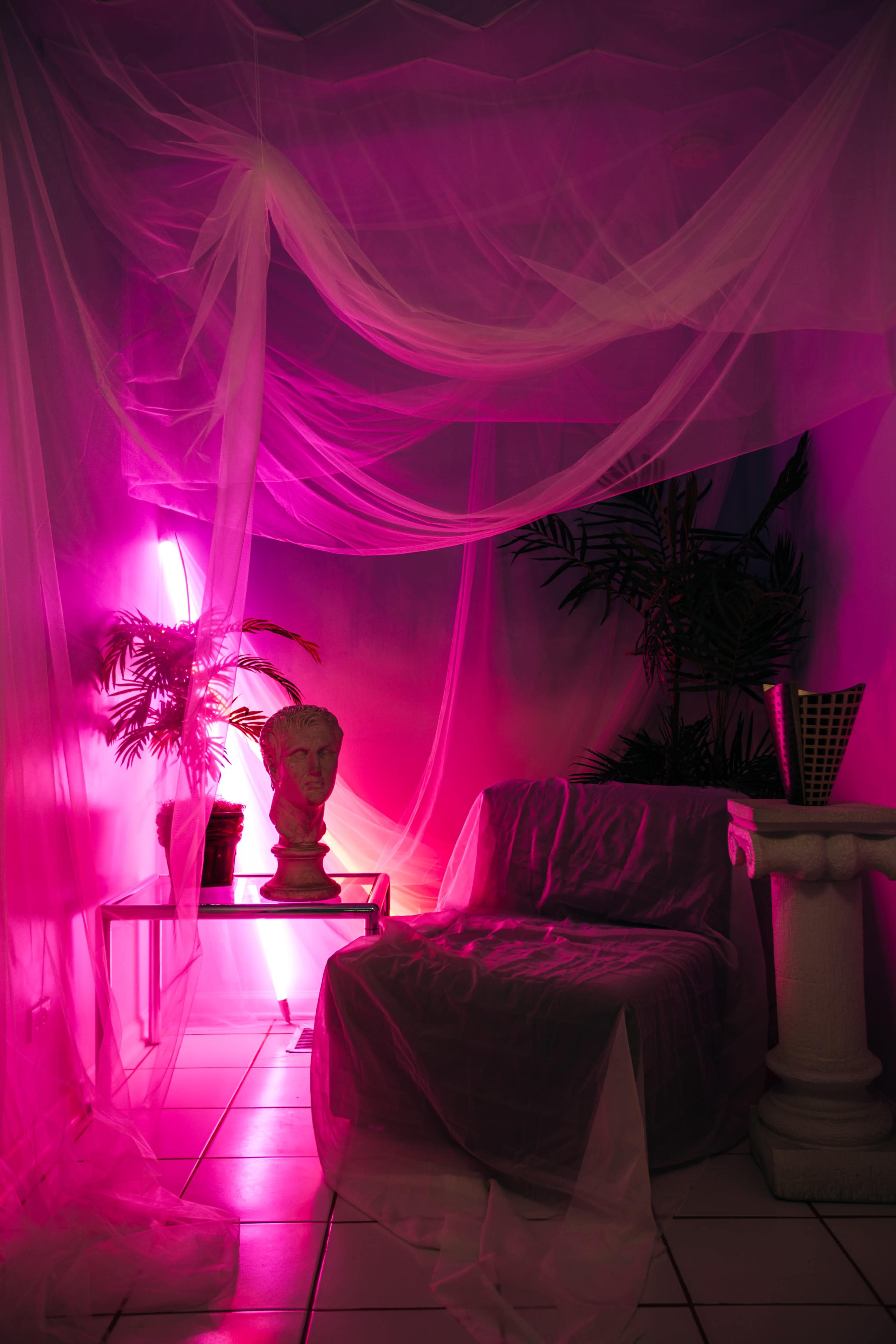 A dimly lit room features a transparent drapery setup, a bust on a pedestal, a small table with a plant, and a chair covered in fabric.