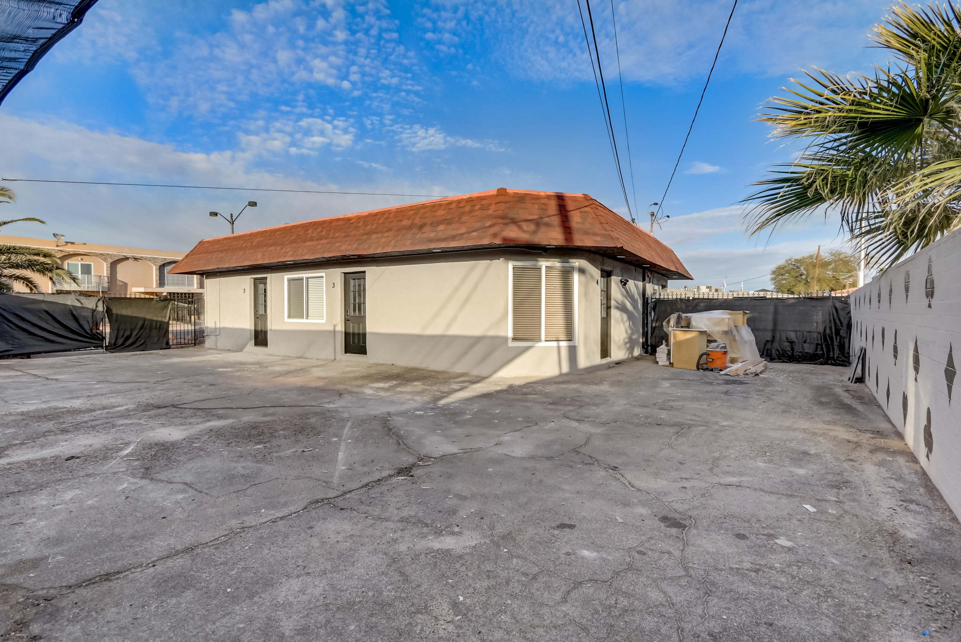 The image shows a one-story building with a red-tiled roof situated on a concrete lot, surrounded by a fence and some palm trees.