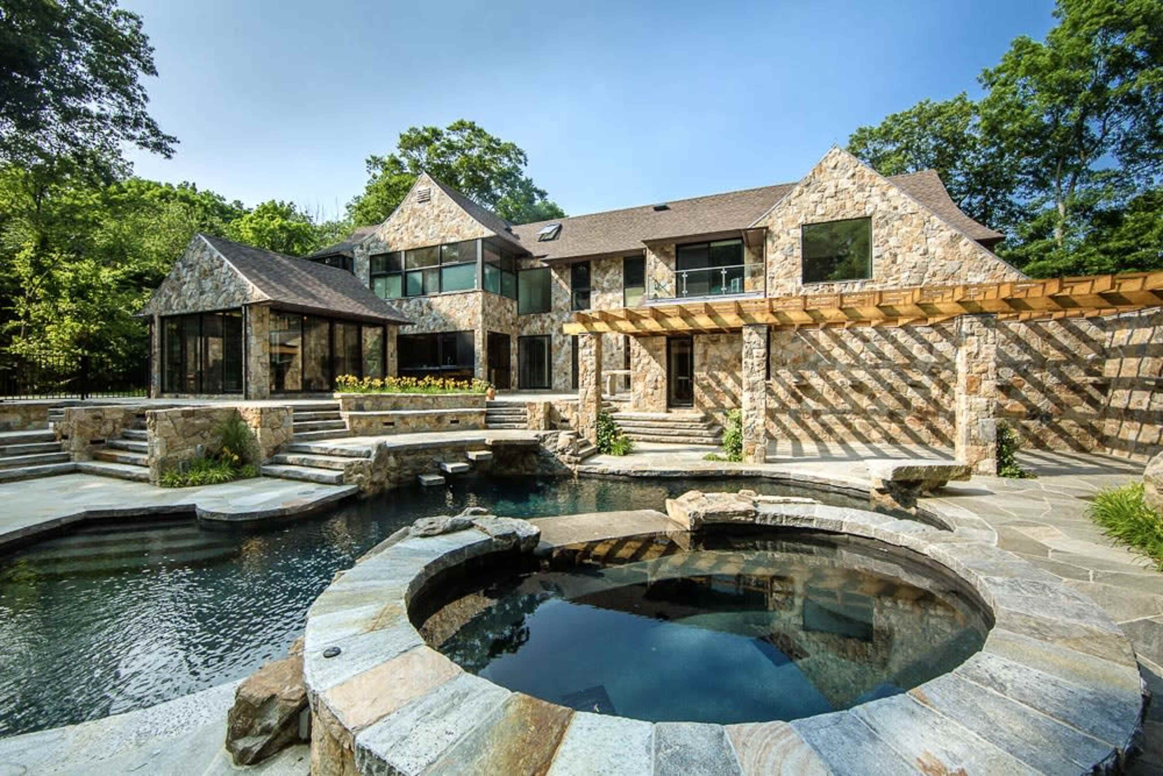 The image shows a large, stone-clad house with multiple windows, a pool, and a deck area surrounded by trees.