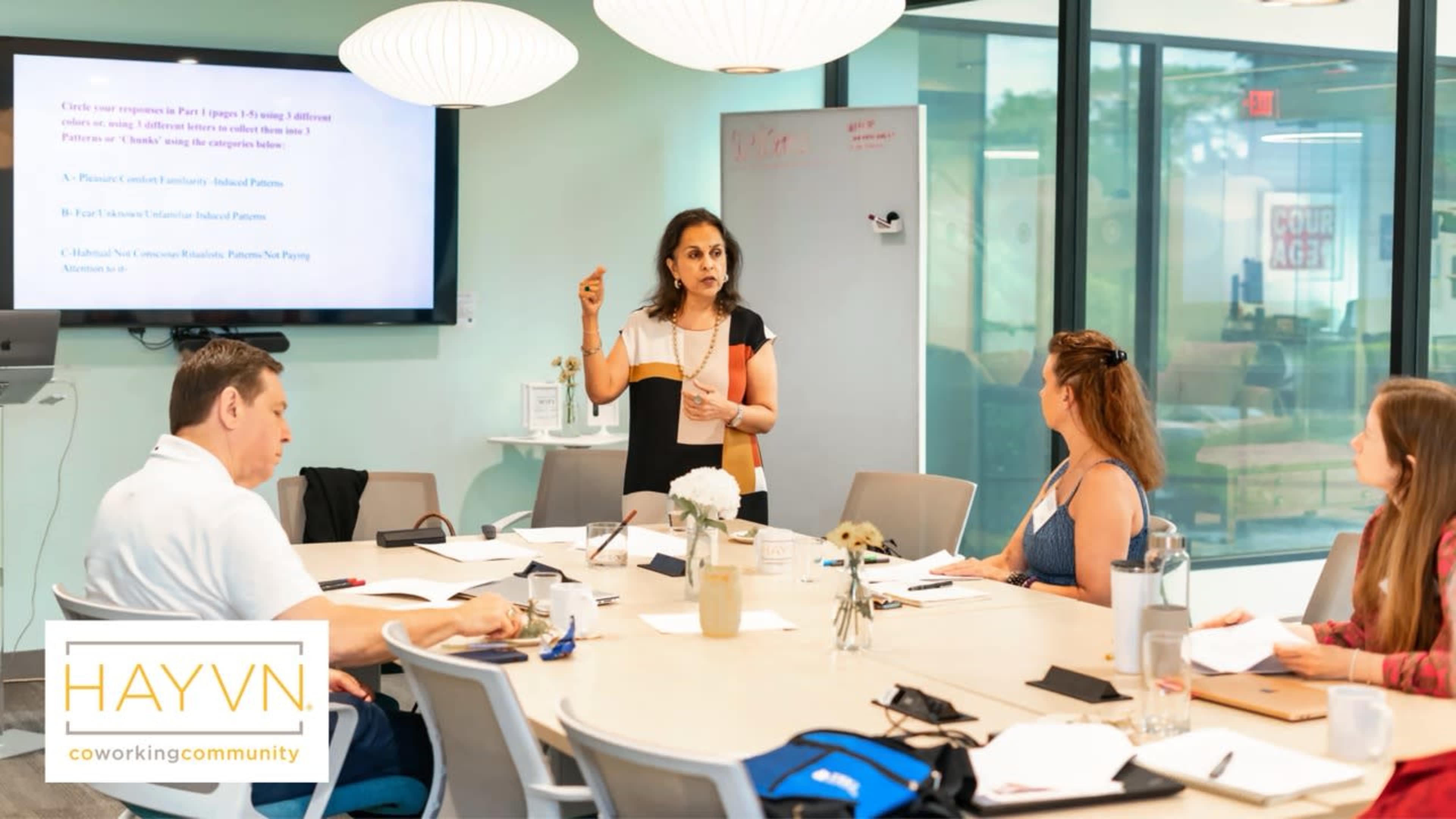 A woman stands at the front of a conference room presenting to a group seated around a table with notebooks and laptops.