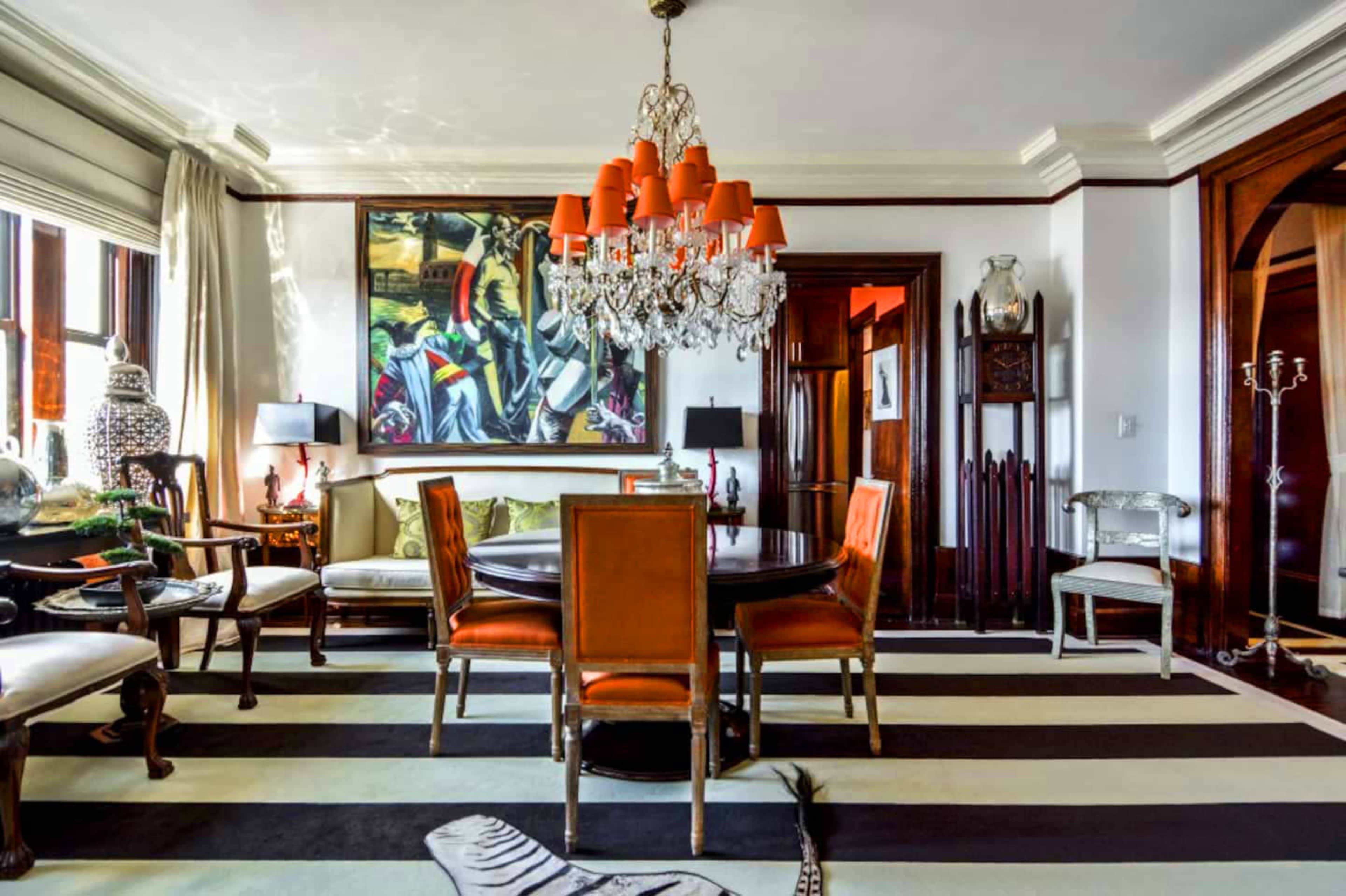 A dining room features a round table surrounded by orange chairs, a crystal chandelier, and artwork on the walls, all set against a striped rug.