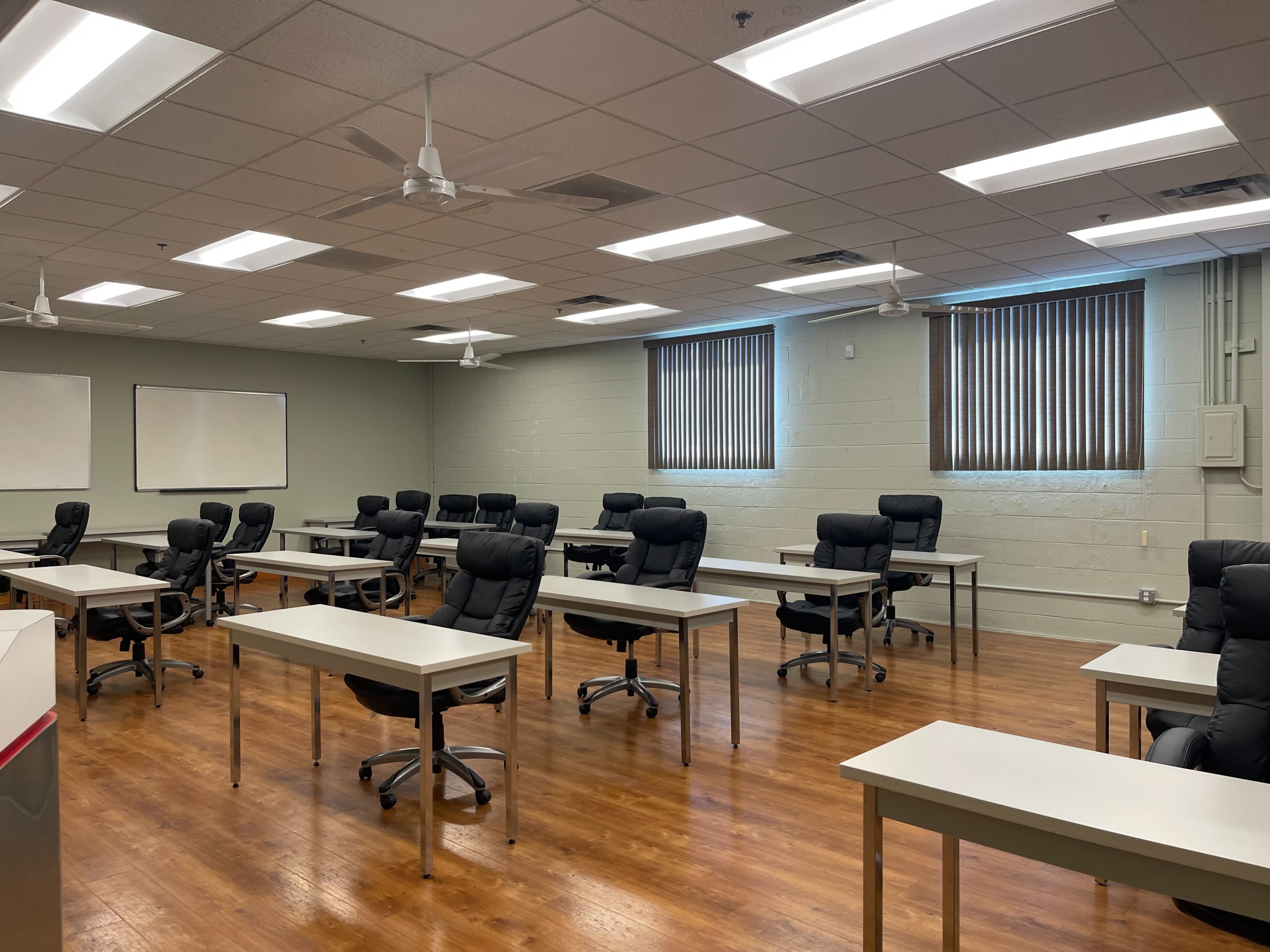 The image shows a classroom with several desks and chairs organized in rows, featuring large windows that let in natural light.