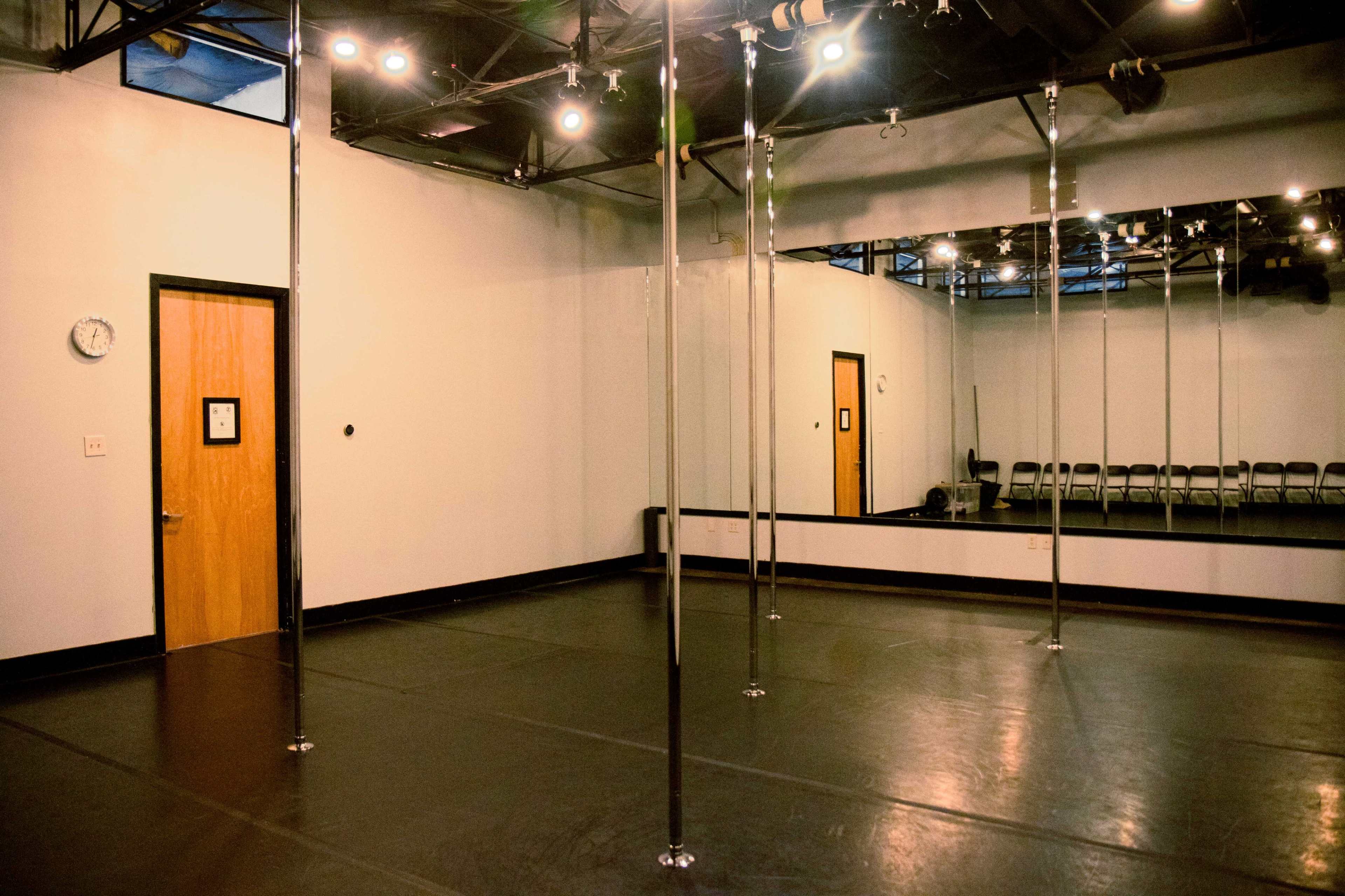 The image shows an empty dance studio with several pole structures, a mirrored wall, and wooden chairs lining one side.
