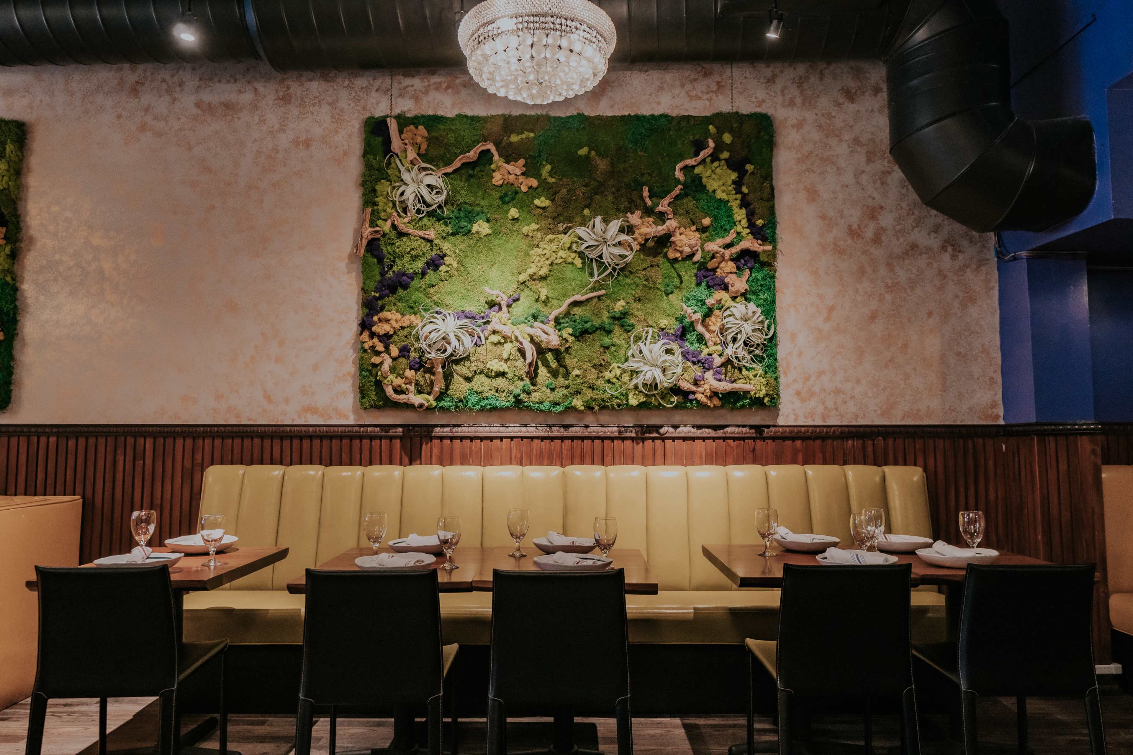 The image shows a modern restaurant interior featuring a yellow upholstered seating area, a wooden table set for dining, and a large, vibrant green wall decoration made of plants and flowers.