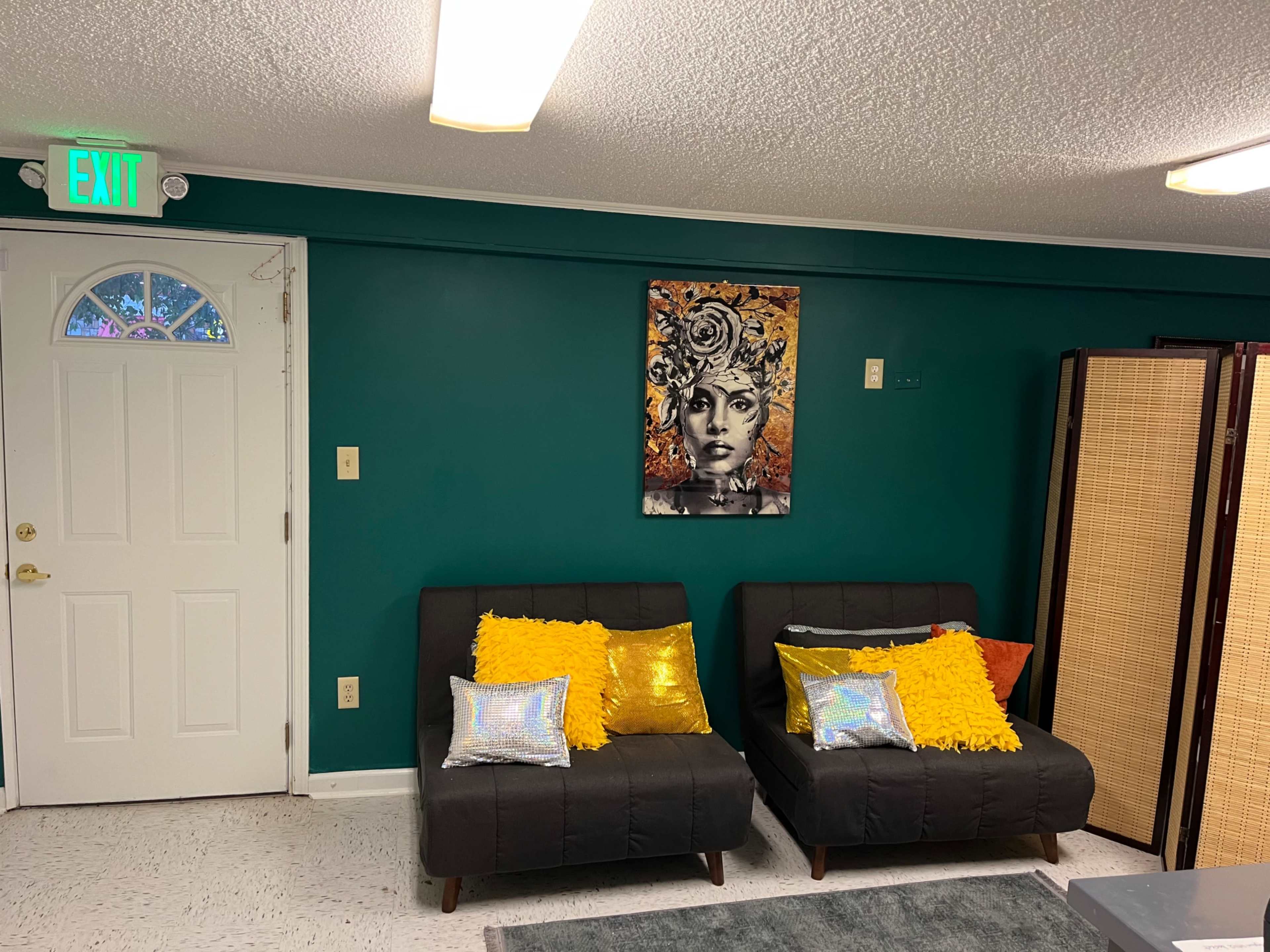 The image shows a cozy living space with two dark-colored sofas adorned with colorful cushions, a vibrant artwork on the teal wall, and a door leading outside.