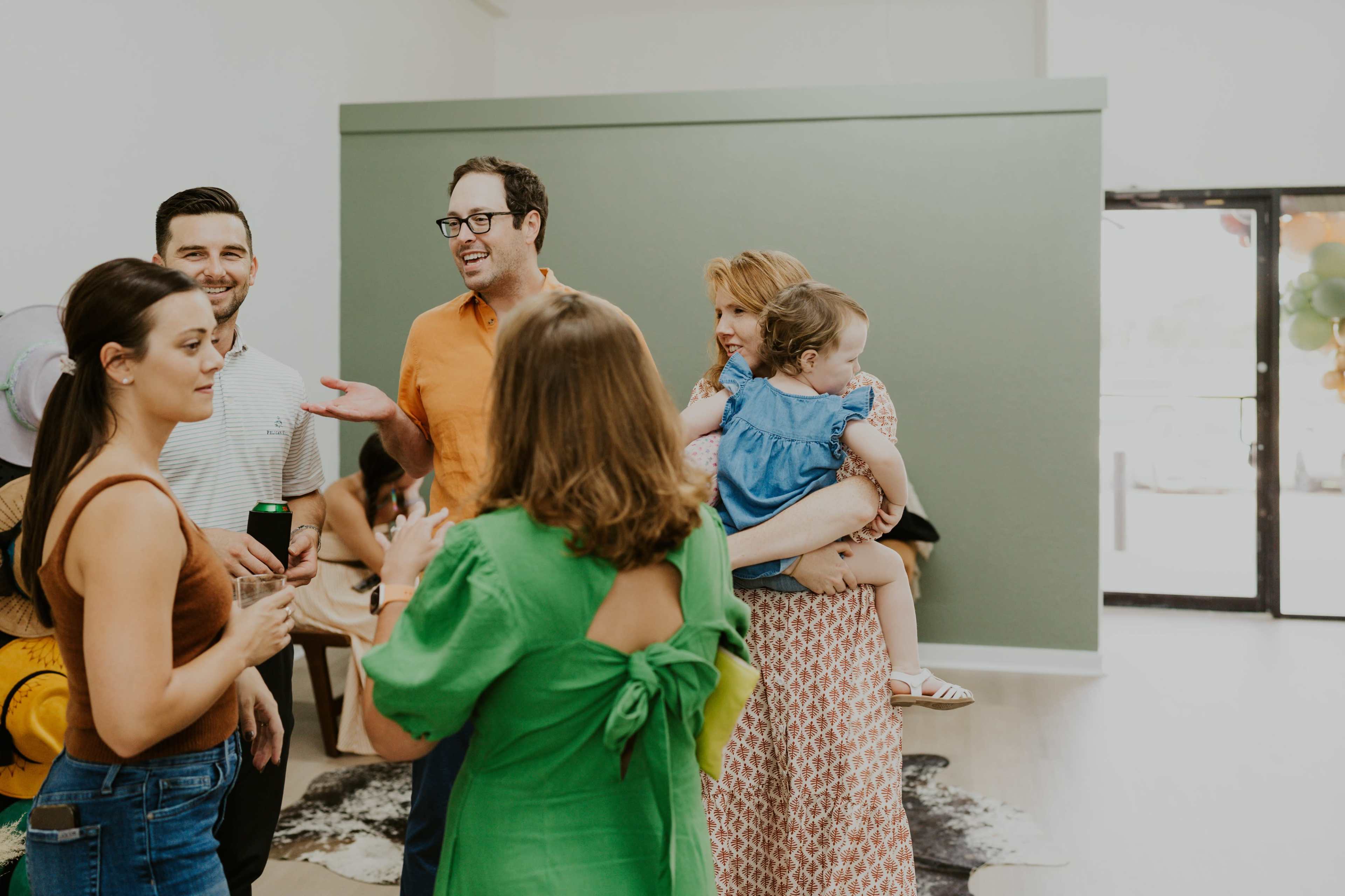 A group of people socializes in a spacious room with green accents, some holding drinks and a child.