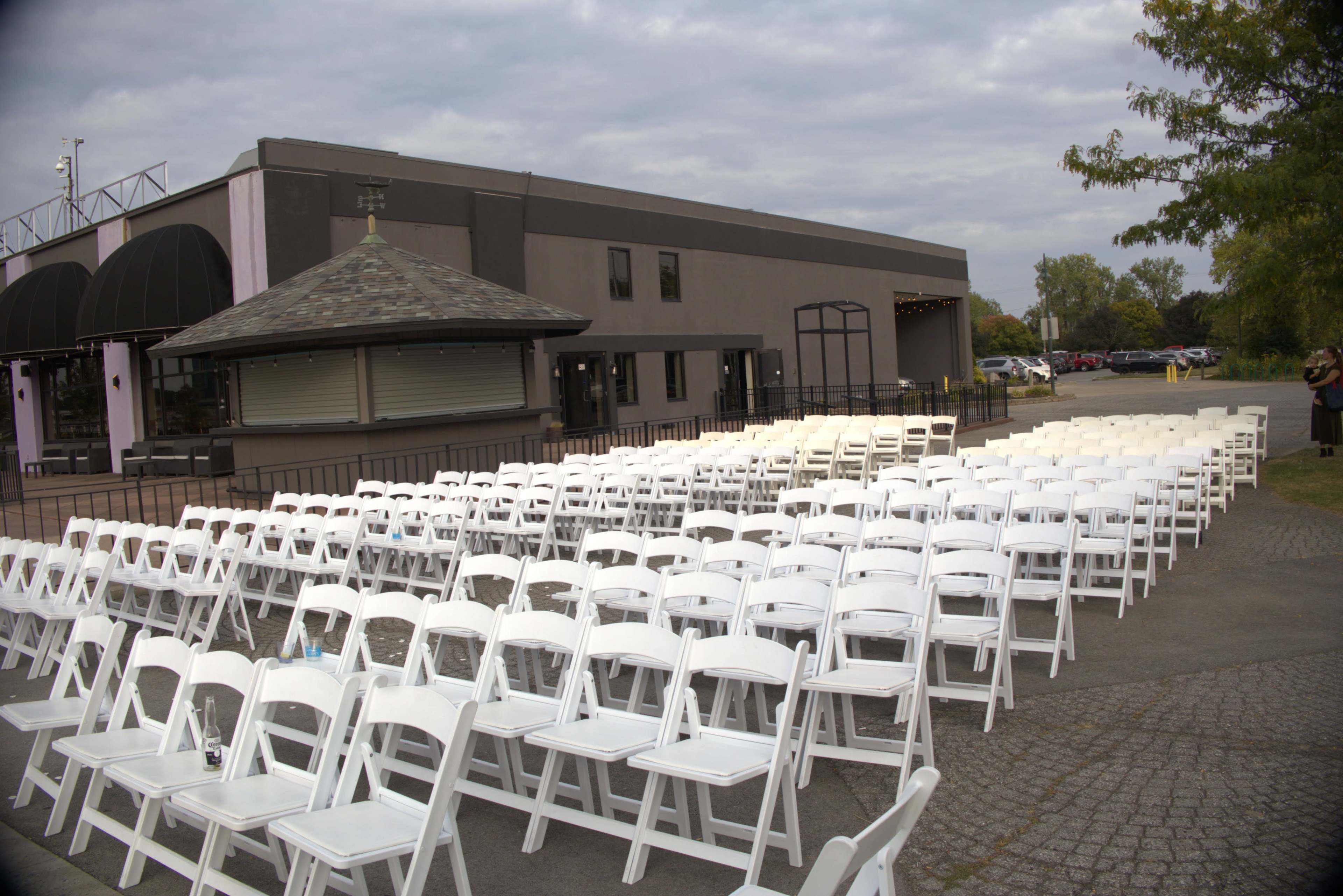 The image shows a large outdoor seating area with rows of white chairs arranged in front of a building under an overcast sky.
