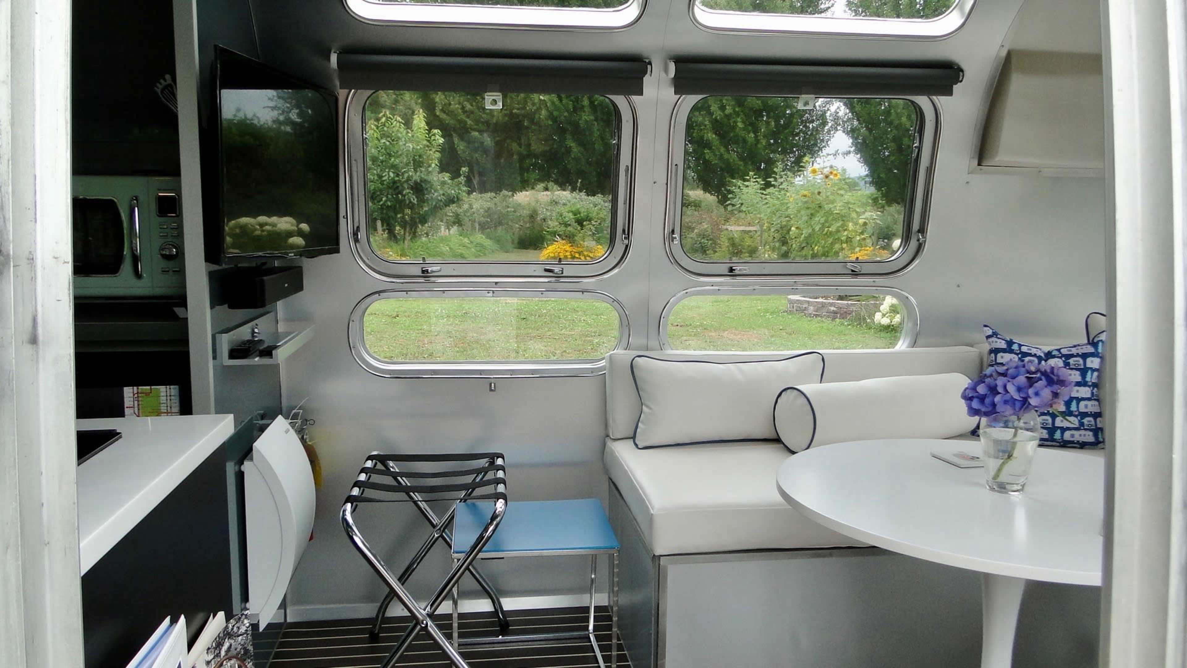 The image shows the interior of a camper with a circular table, a couch, and large windows that overlook a garden.