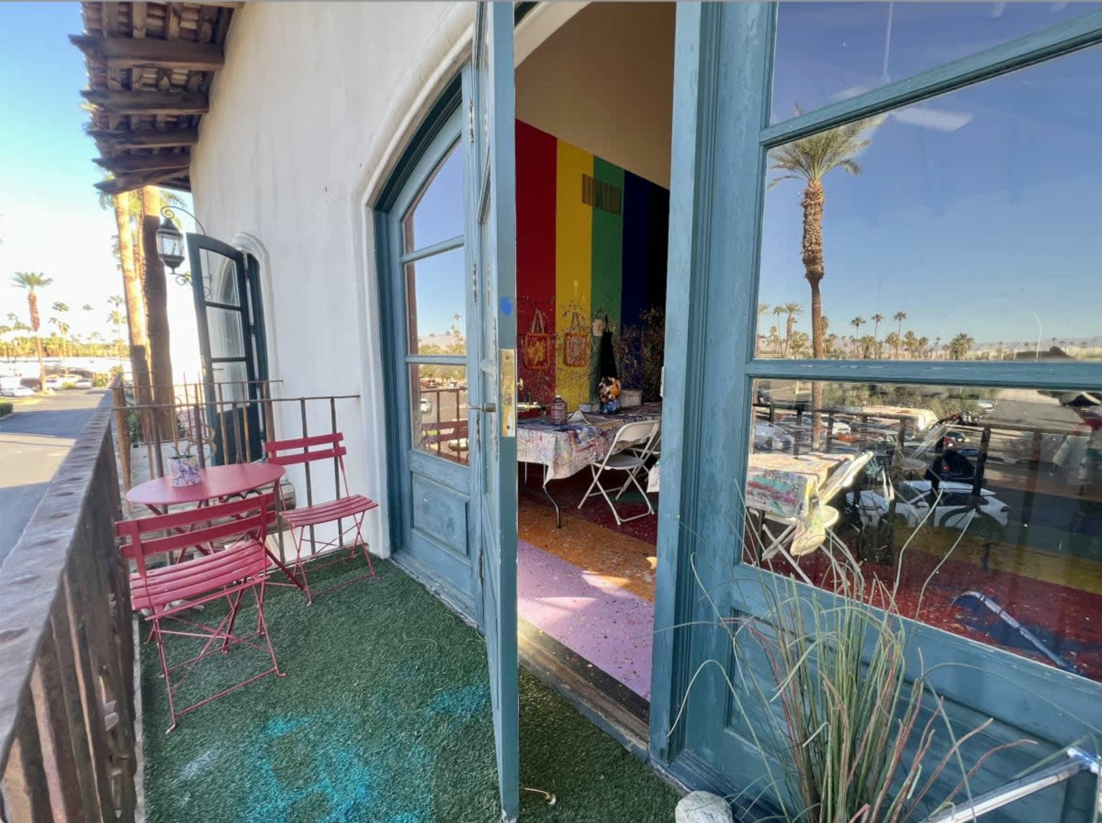 The image shows an open doorway leading to a colorful interior space, with outdoor seating on a patio overlooking palm trees.