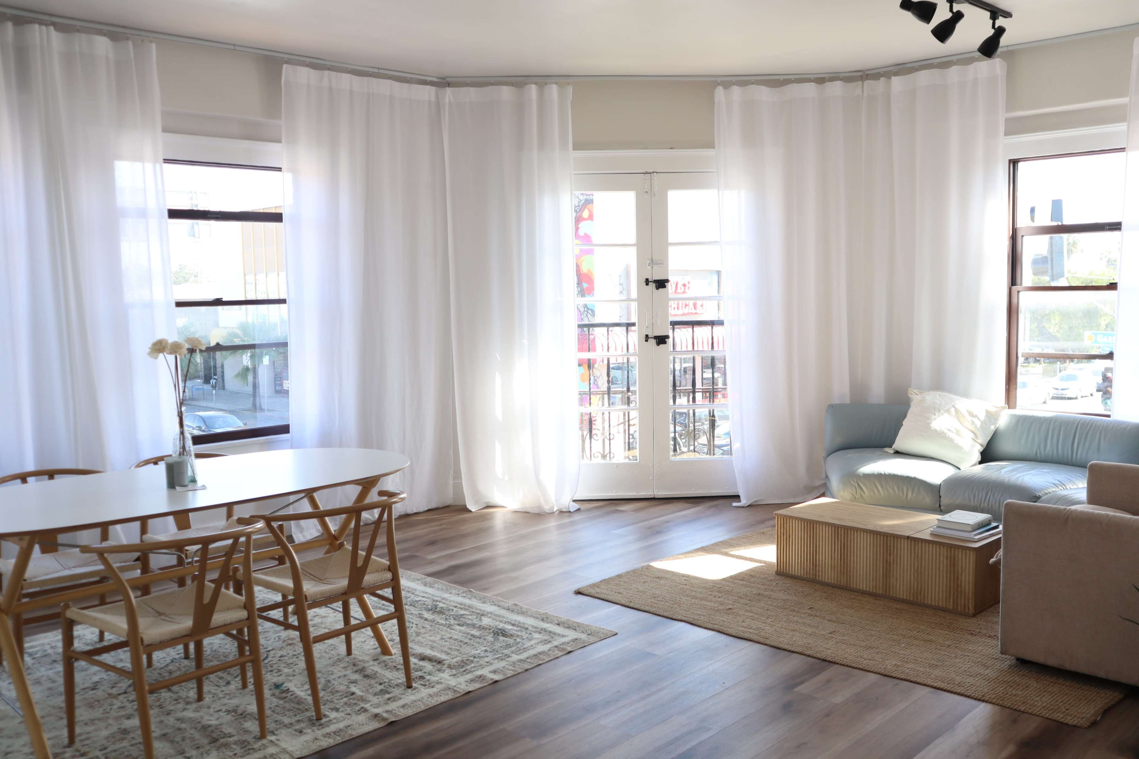 The image shows a modern, well-lit living space featuring a round dining table, light-colored curtains, a blue sofa, and natural wood accents.