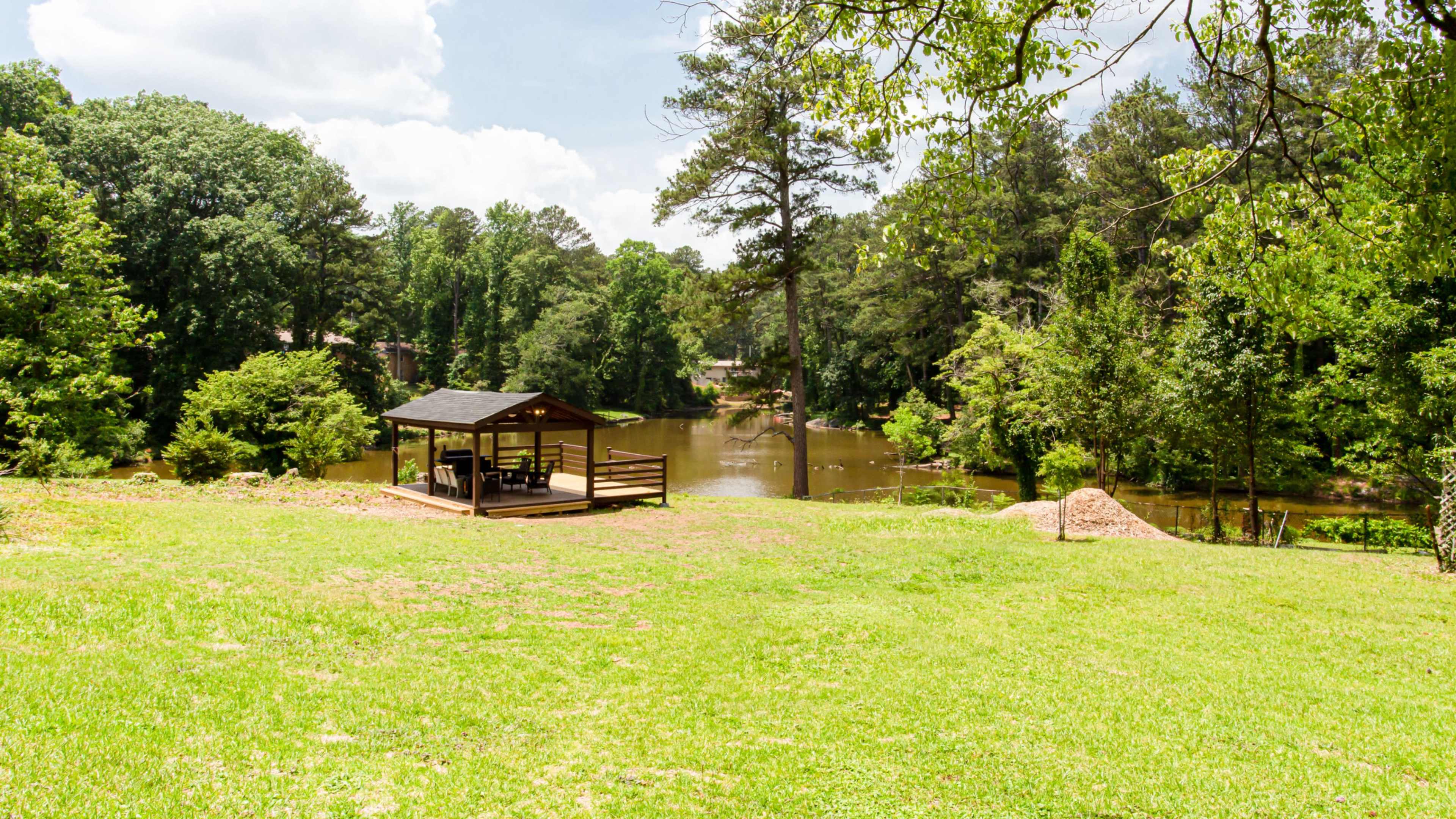 A wooden gazebo sits on a grassy area overlooking a calm pond surrounded by trees.