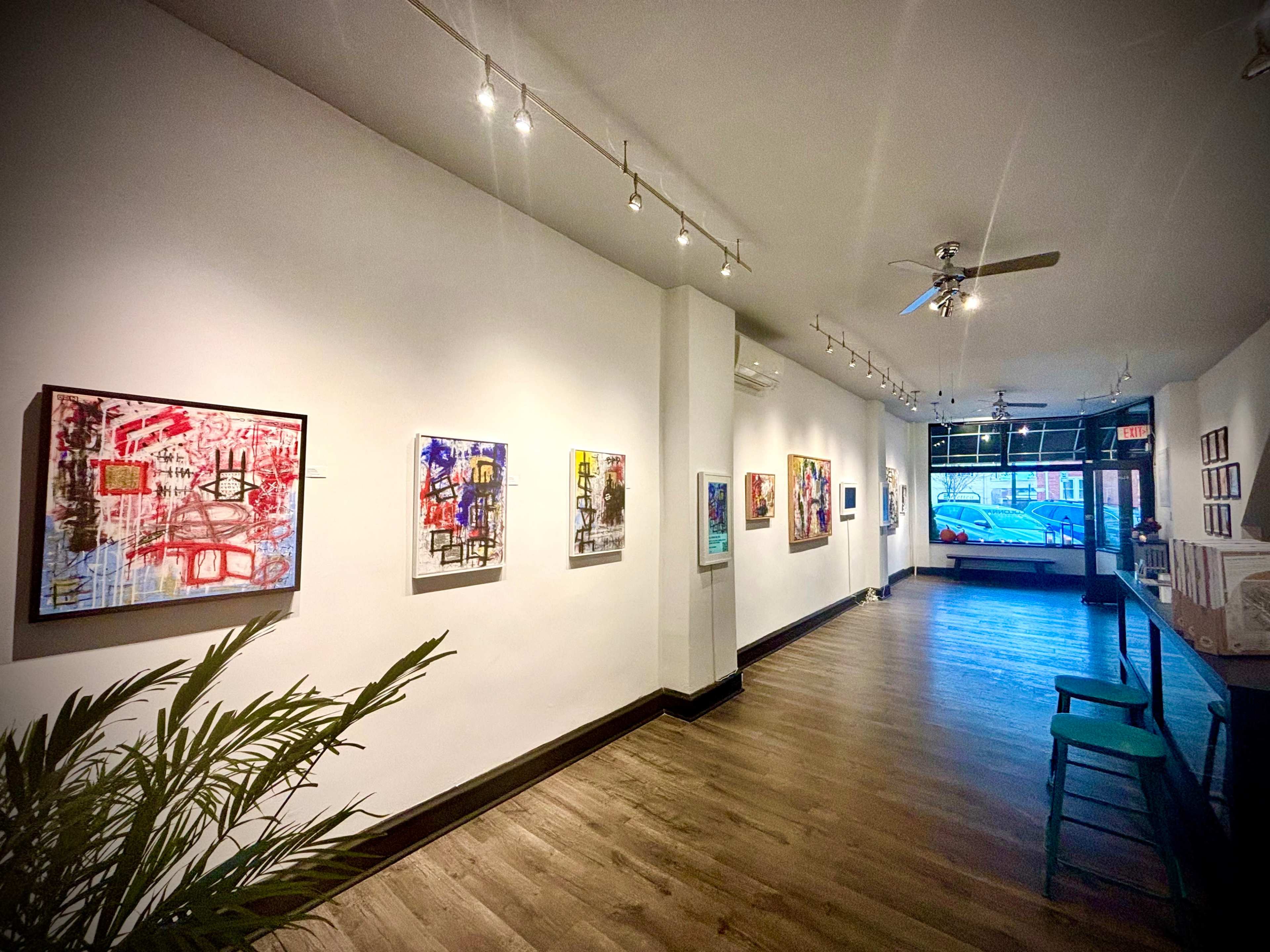 A gallery interior with artwork displayed on white walls and wooden flooring, featuring a row of paintings on one side and a seating area on the left.