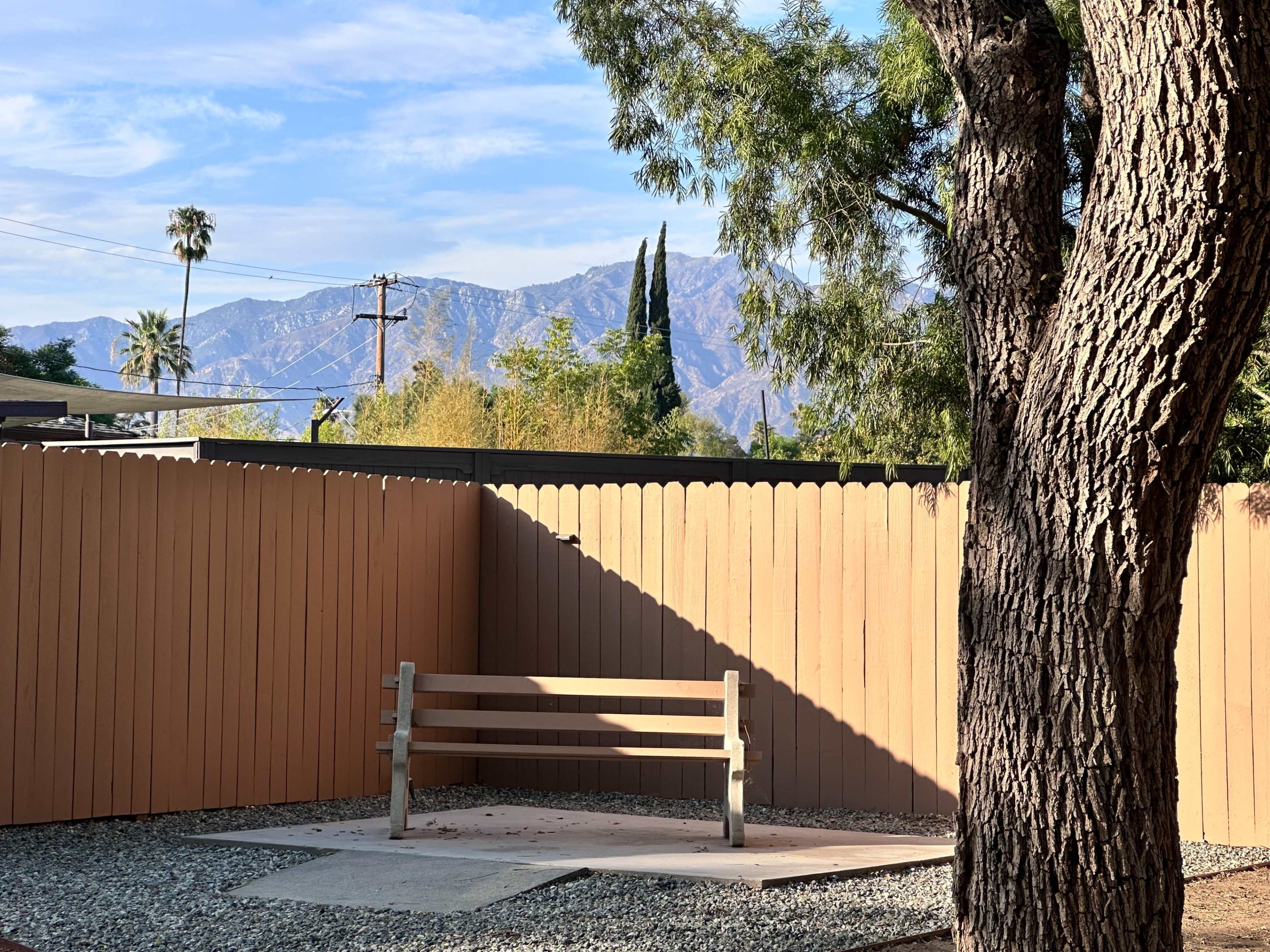 A wooden bench sits on a gravel surface against a backdrop of mountains and a clear blue sky, enclosed by a wooden fence.