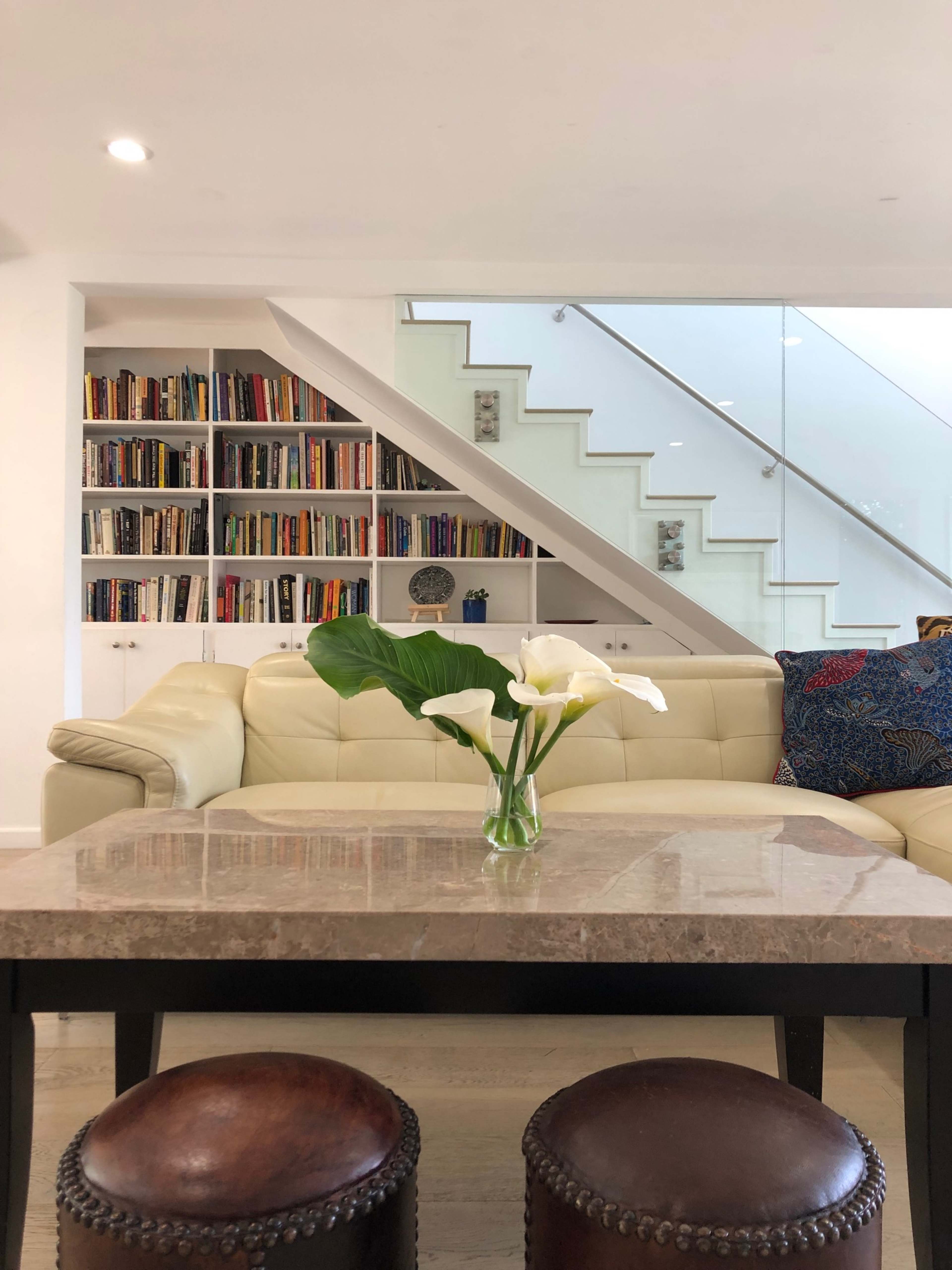 A modern living room features a beige sofa, a glass staircase, a bookshelf filled with books, and a coffee table with a vase of white flowers.