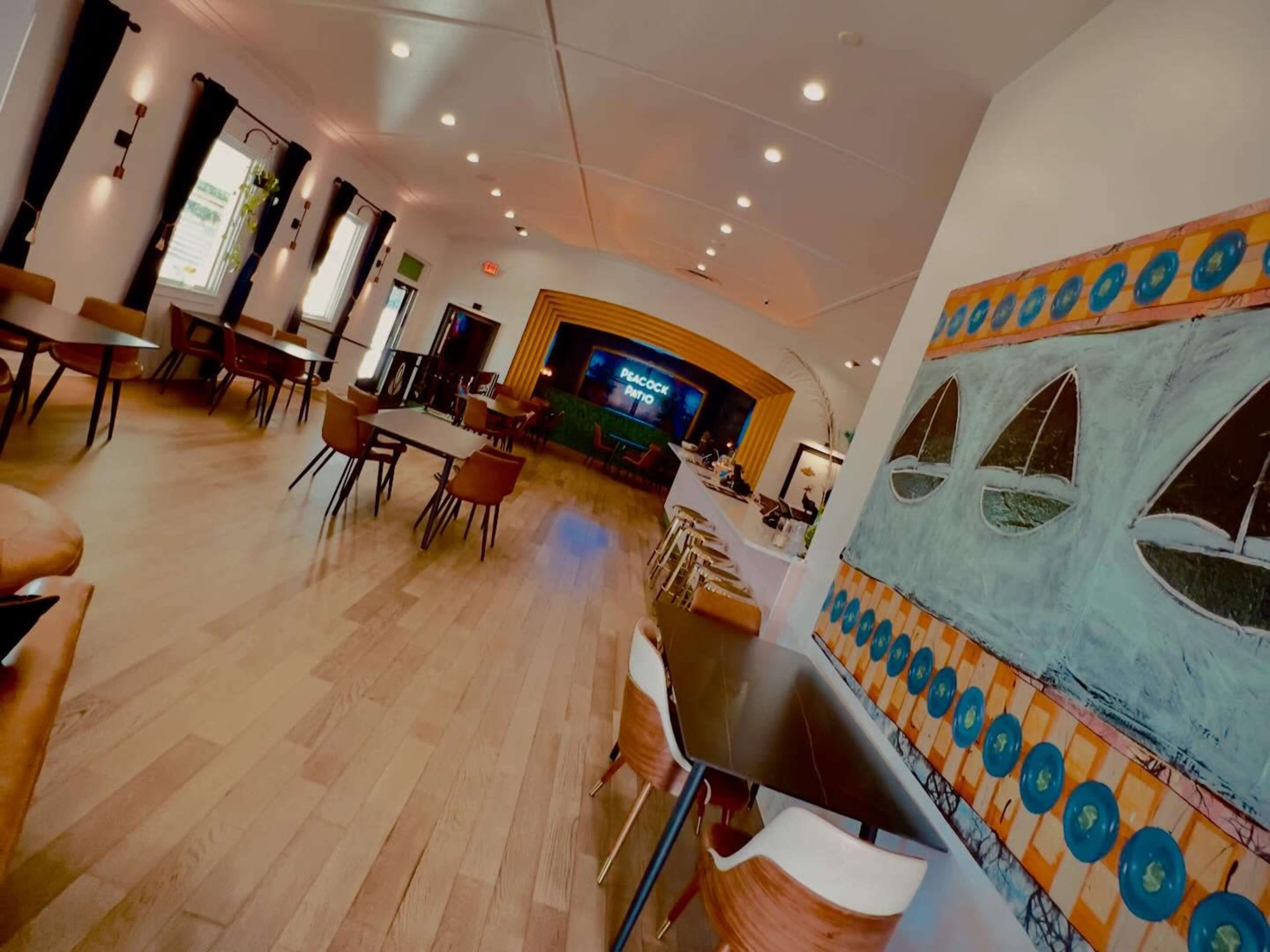 The image shows a spacious interior of a modern café with wooden flooring, a bar area, several tables and chairs, and a large mural featuring boats on the wall.