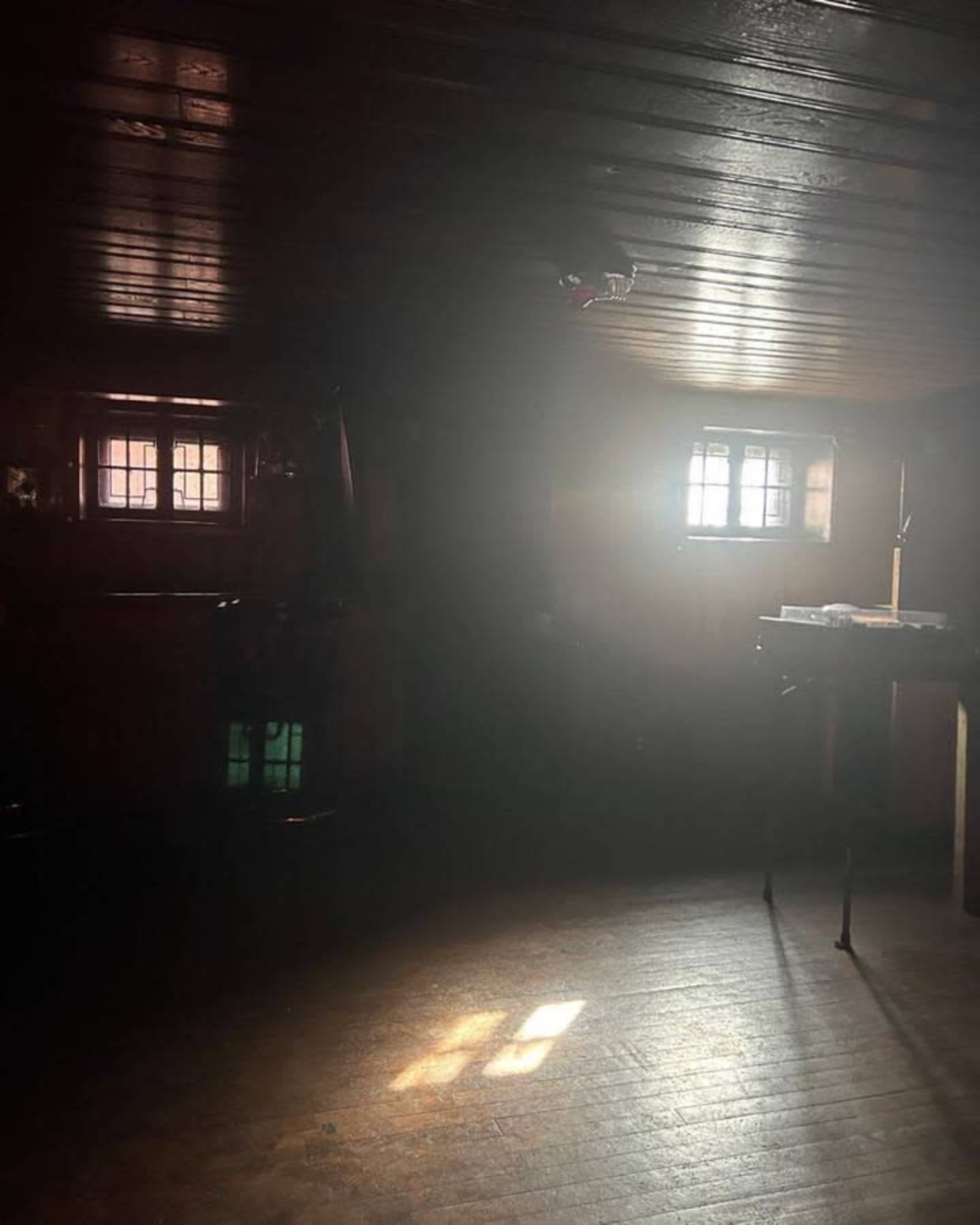 A dimly lit room features beams of light streaming through two small windows onto the wooden floor.