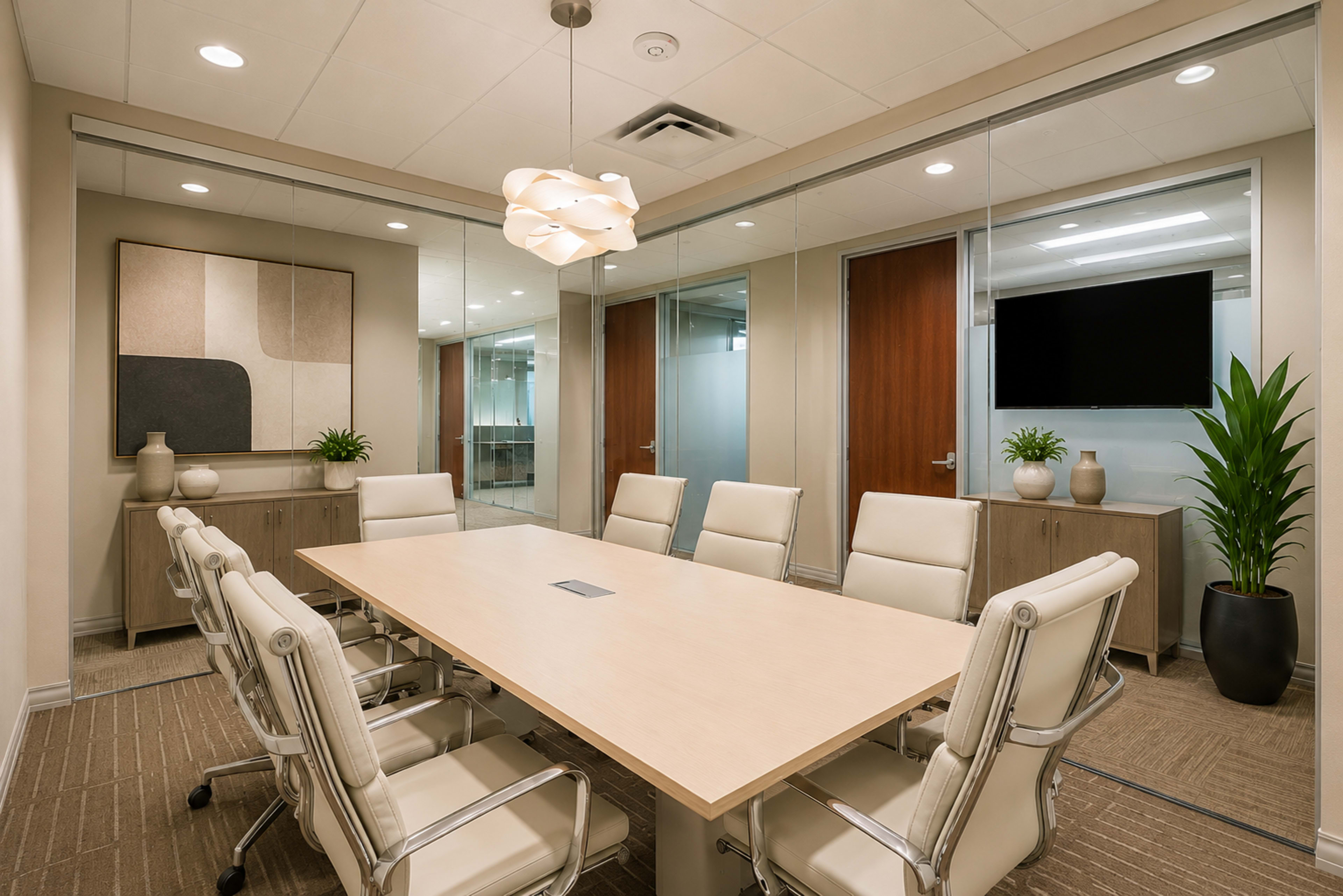 A modern conference room features a long wooden table surrounded by ergonomic white chairs, with glass walls, decorative plants, and a mounted television.