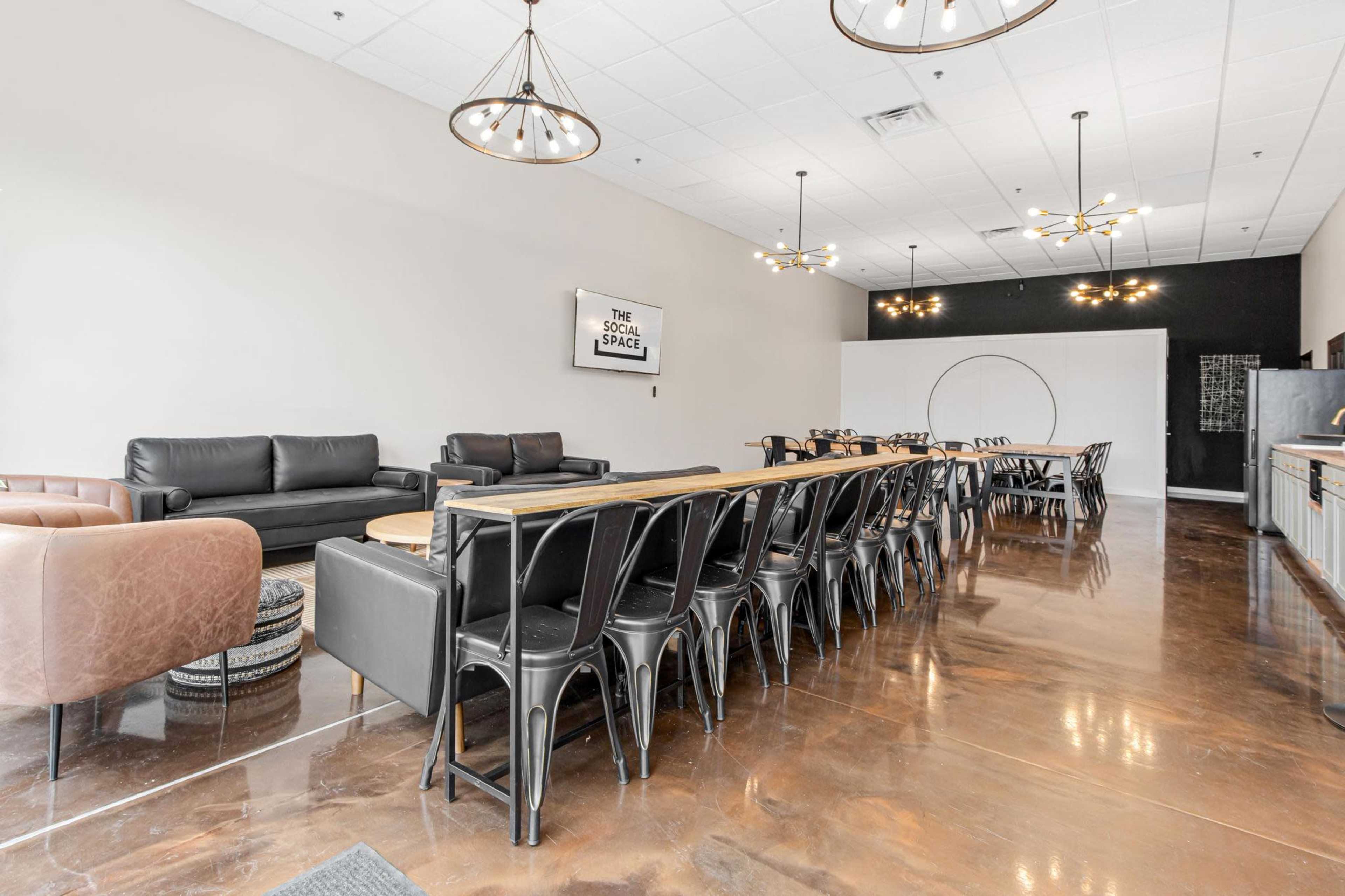 A spacious, modern meeting area featuring a long wooden table surrounded by metal chairs, leather sofas, and contemporary lighting fixtures.