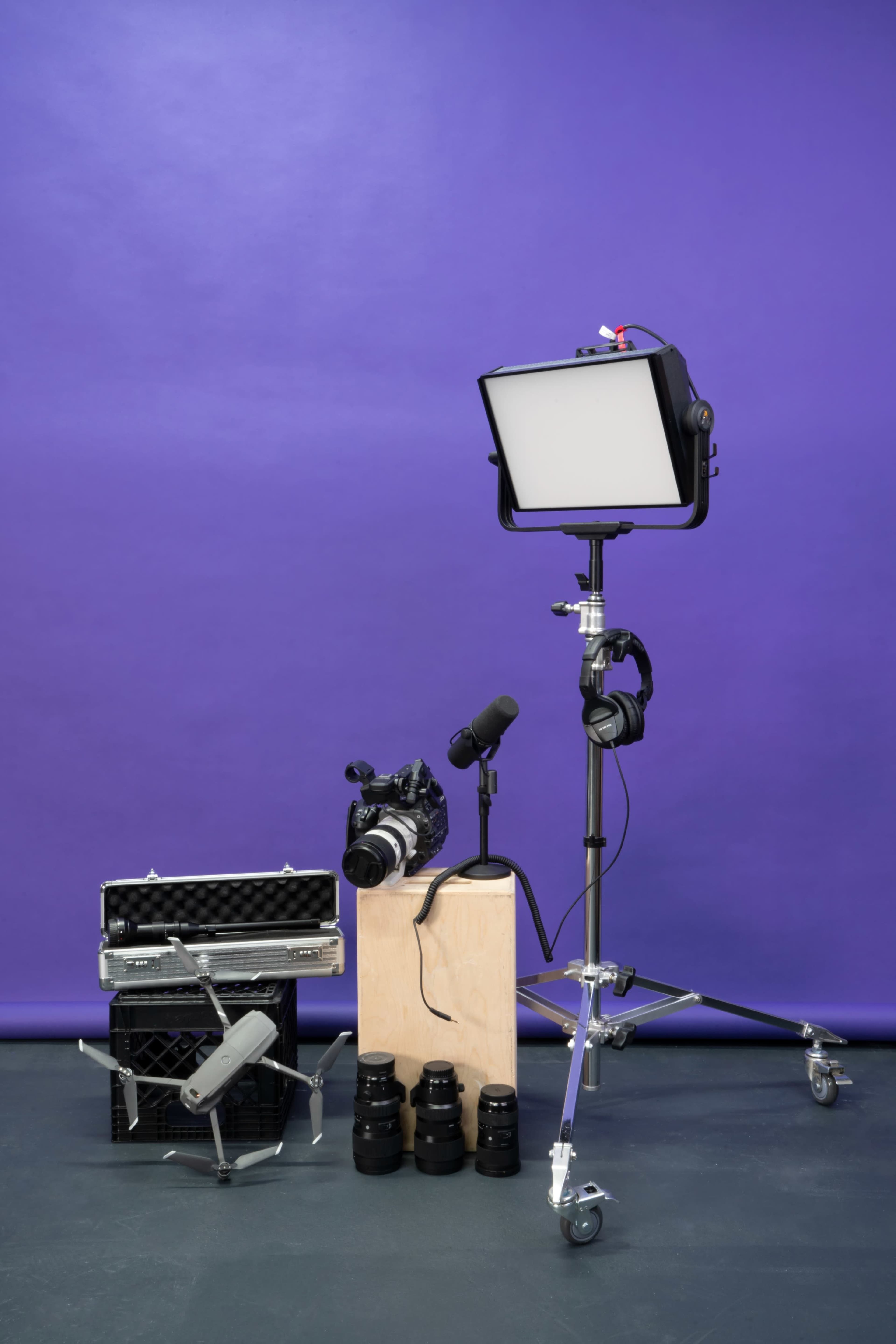 The image features a photography setup with a light on a stand, a camera, microphones, headphones, various camera lenses, and a drone arranged against a purple backdrop.