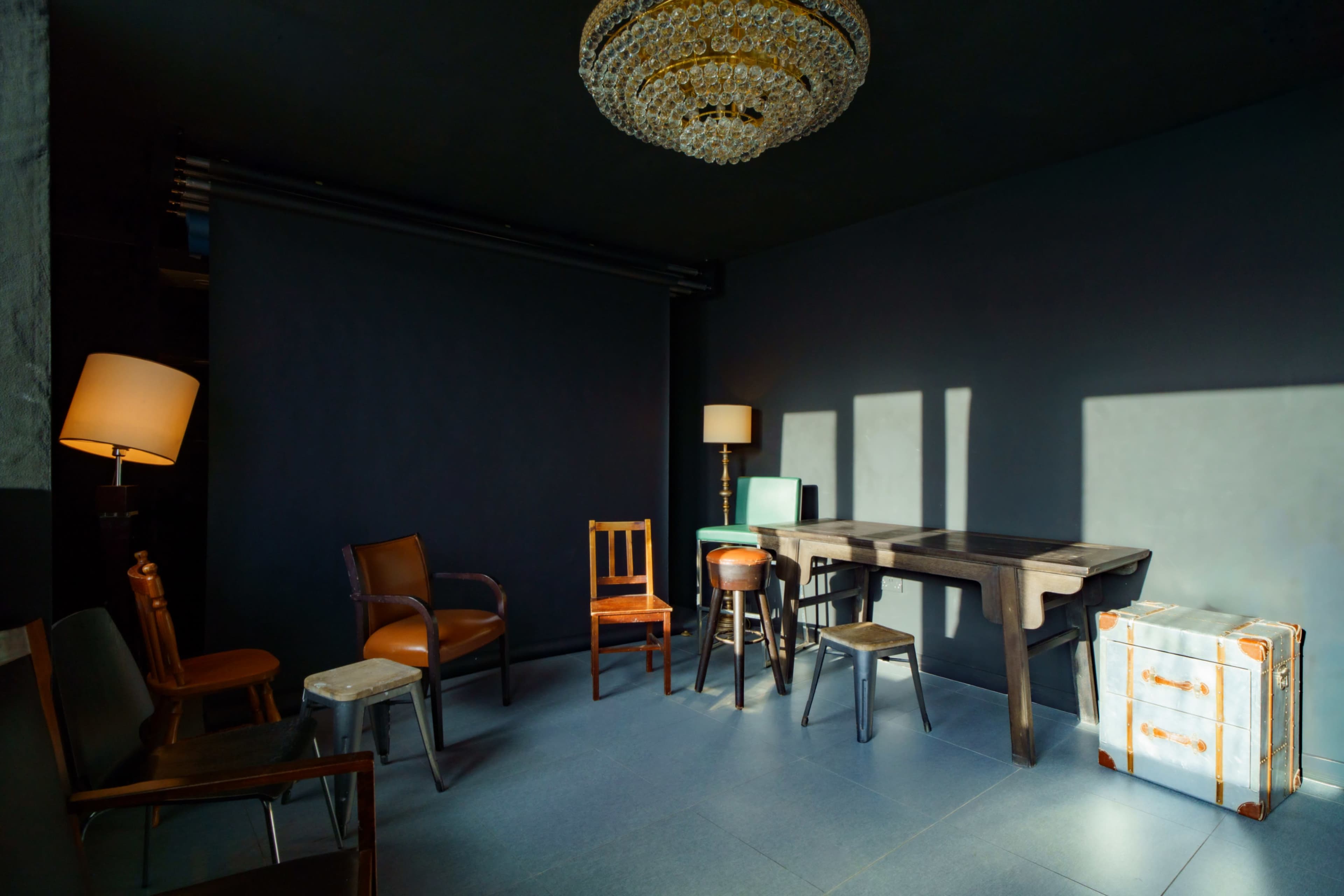 A well-lit room features a variety of furniture, including chairs, a table, and a vintage suitcase, against a dark backdrop.
