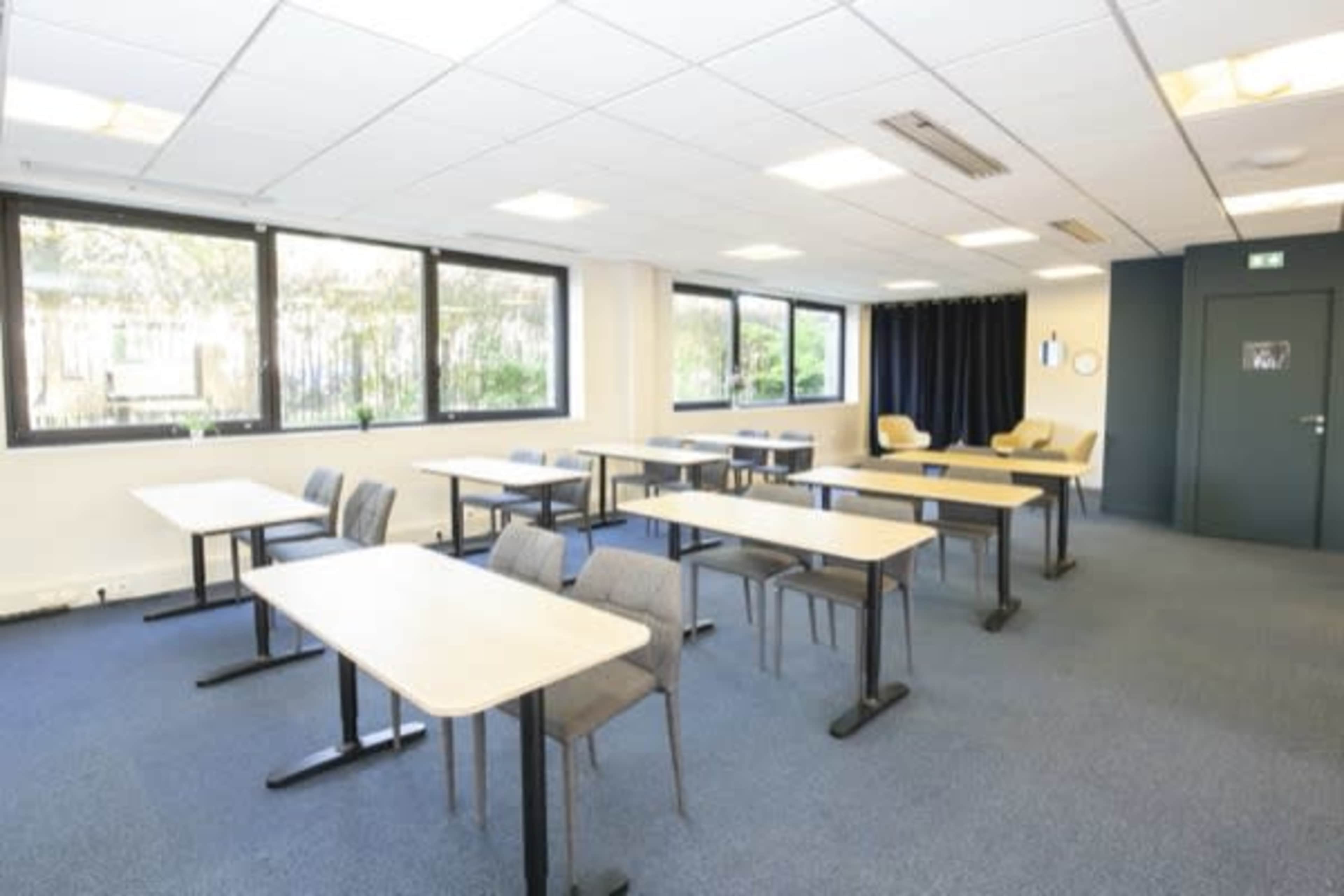 A classroom is arranged with several tables and chairs facing the windows, with a blue carpet and a dark curtain on one side.