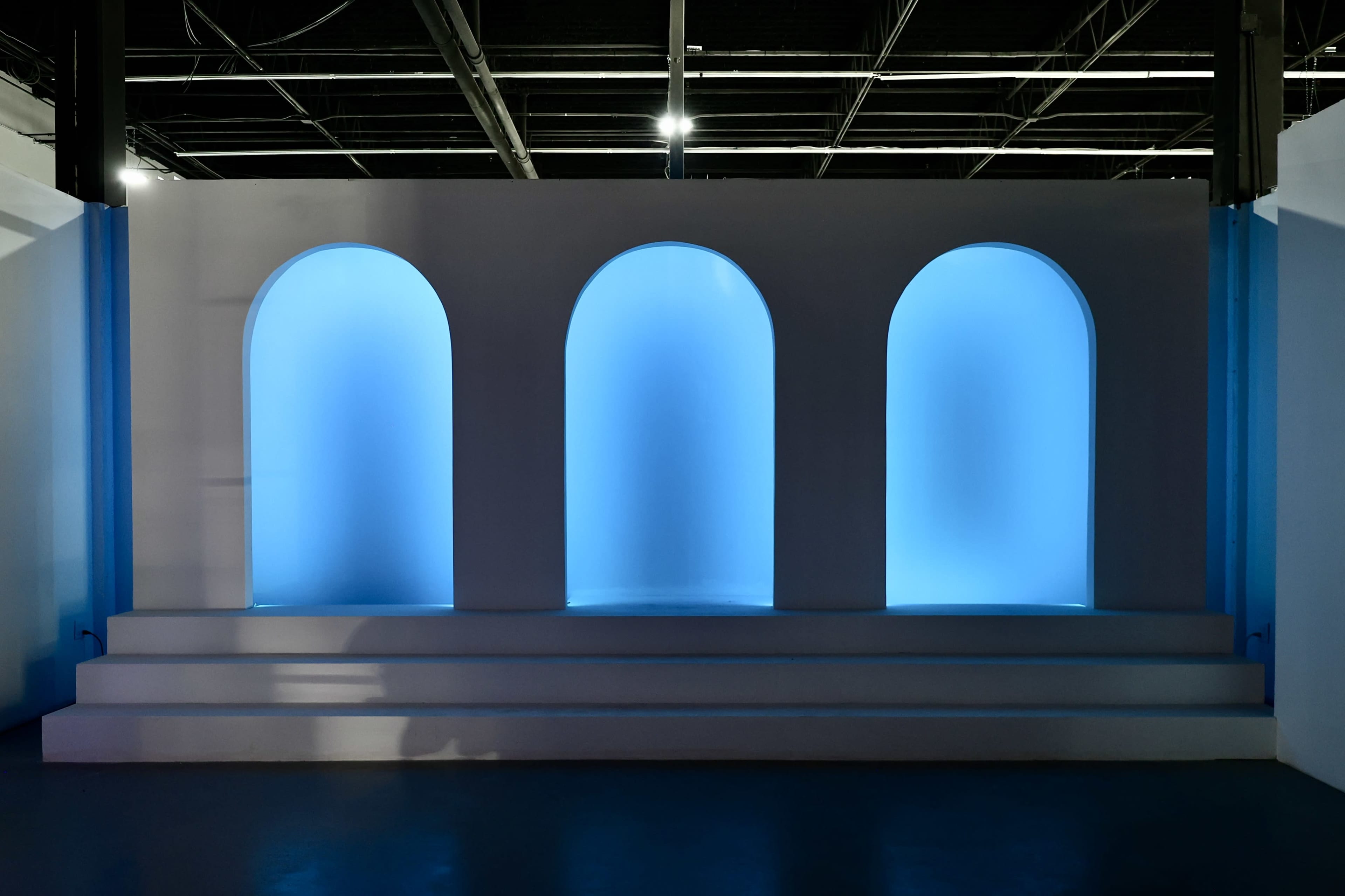 The image shows a minimalist display featuring three arched niches illuminated with a blue light, positioned against a white wall.