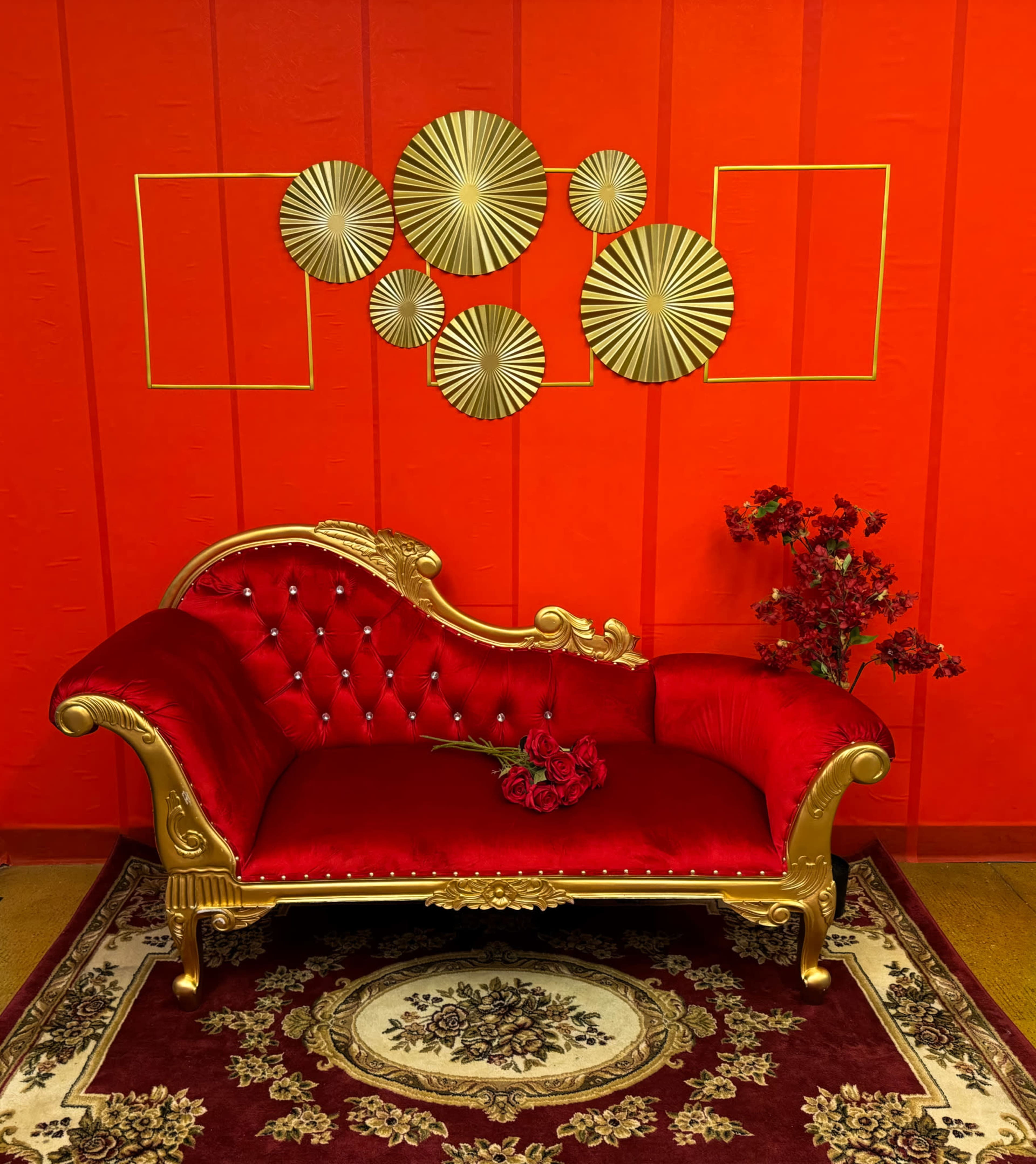 A red velvet couch with gold accents is positioned in front of an orange wall adorned with circular wall decor, accompanied by a floral arrangement and a patterned rug on the floor.