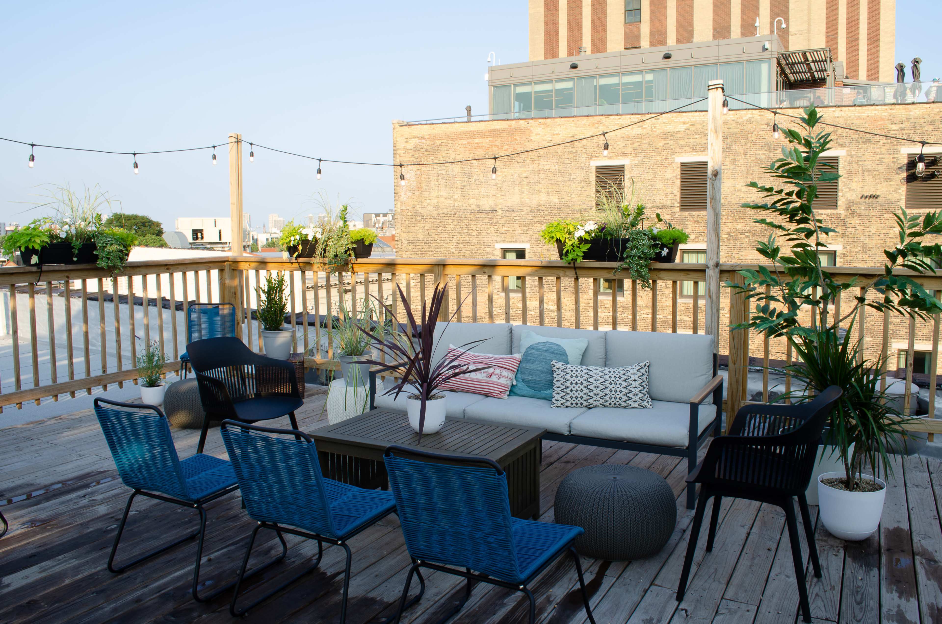 A rooftop space with a seating area that includes a sofa, a coffee table, and several chairs, surrounded by potted plants and string lights overhead.