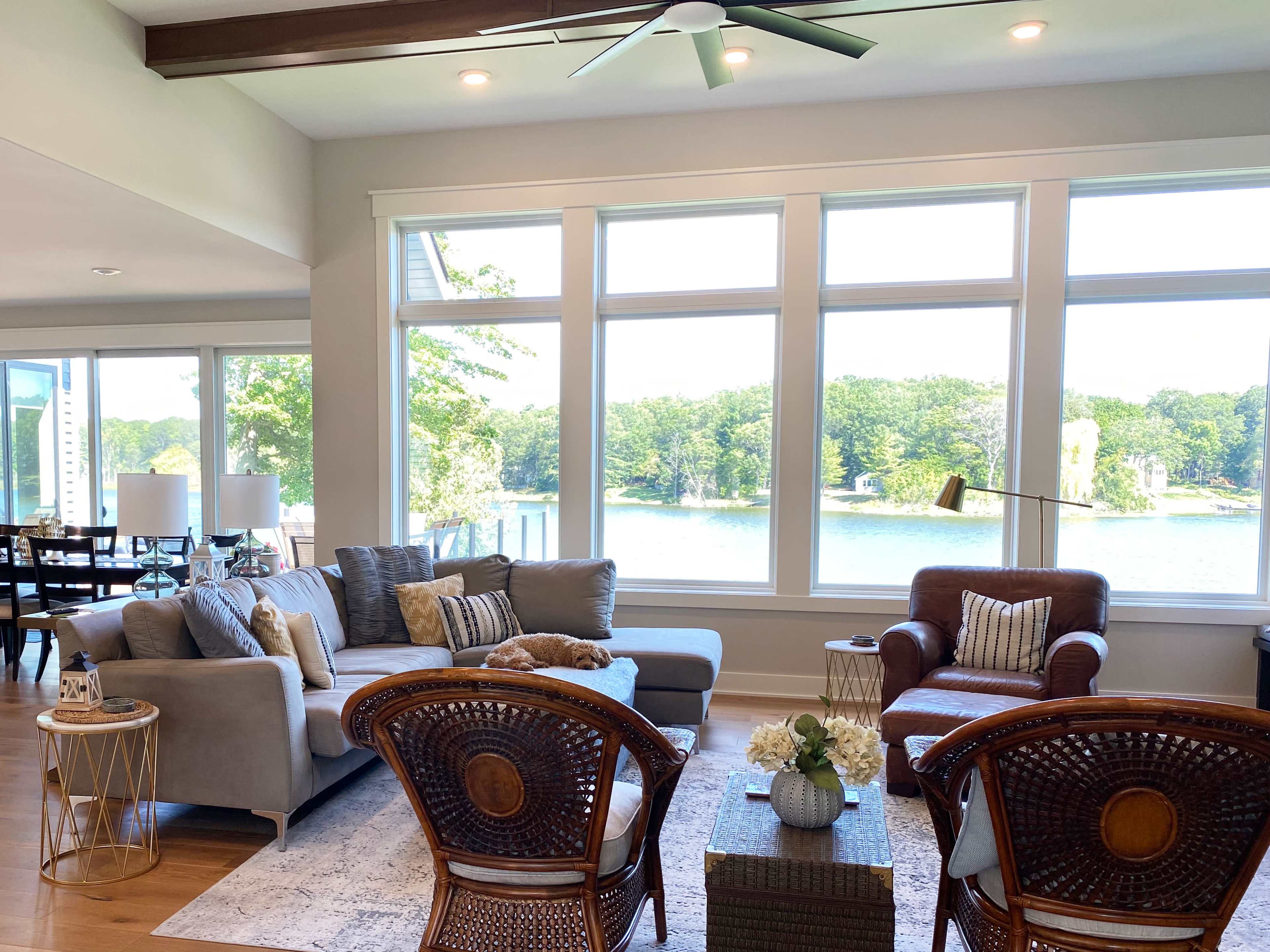 A spacious living room features large windows overlooking a lake, with a gray sofa, a brown armchair, and a wooden coffee table surrounded by woven chairs.