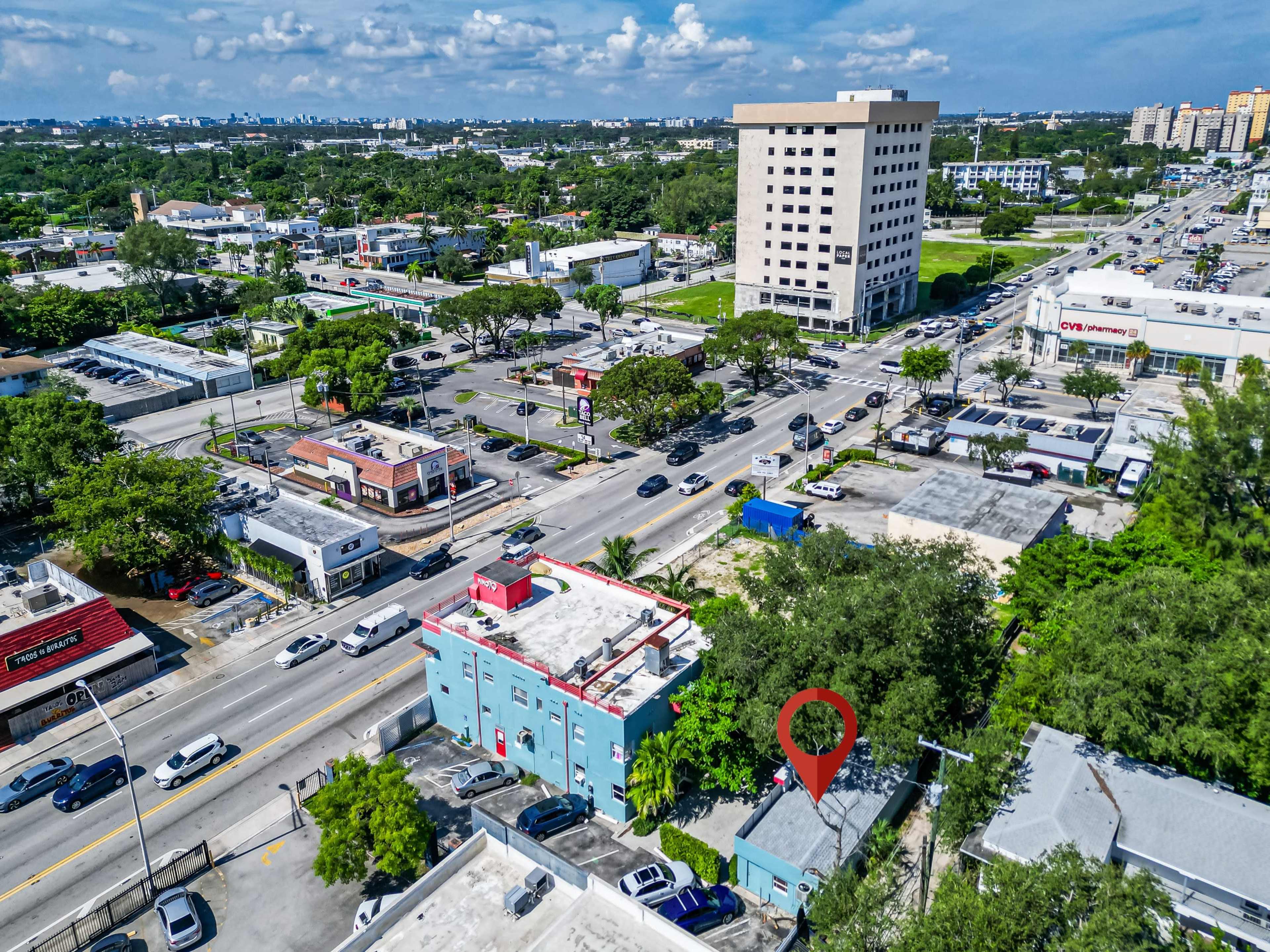 An aerial view shows a bustling urban neighborhood with several buildings, trees, and a busy intersection.