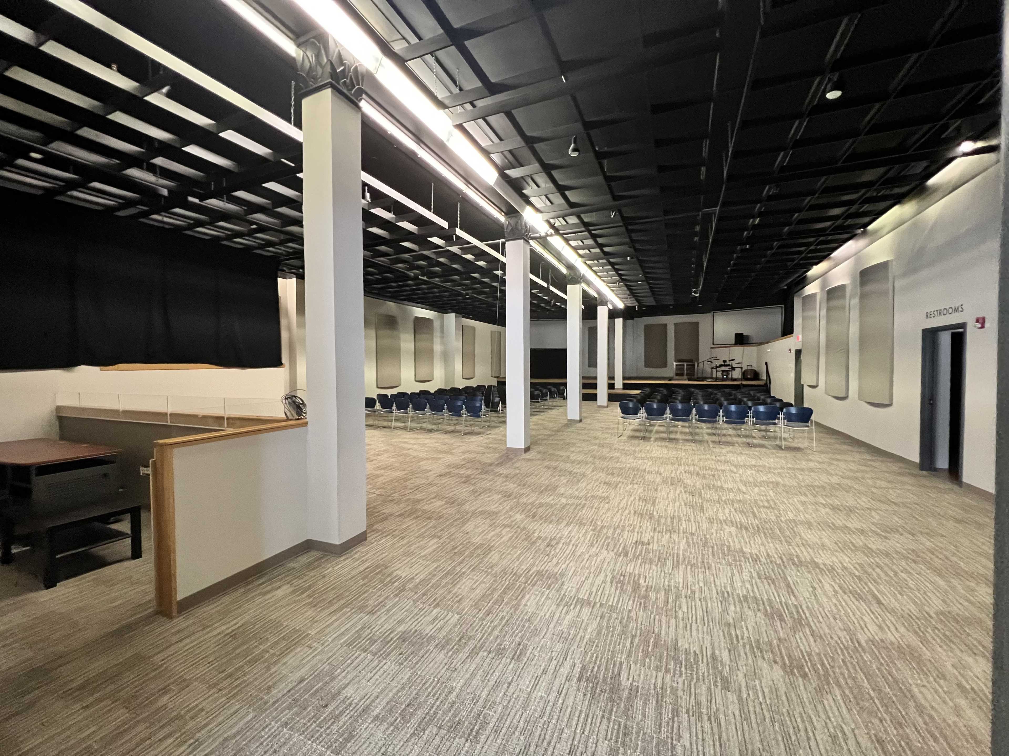 A spacious indoor room with neatly arranged seating, gray walls, and a high ceiling, featuring a stage or platform area at one end.