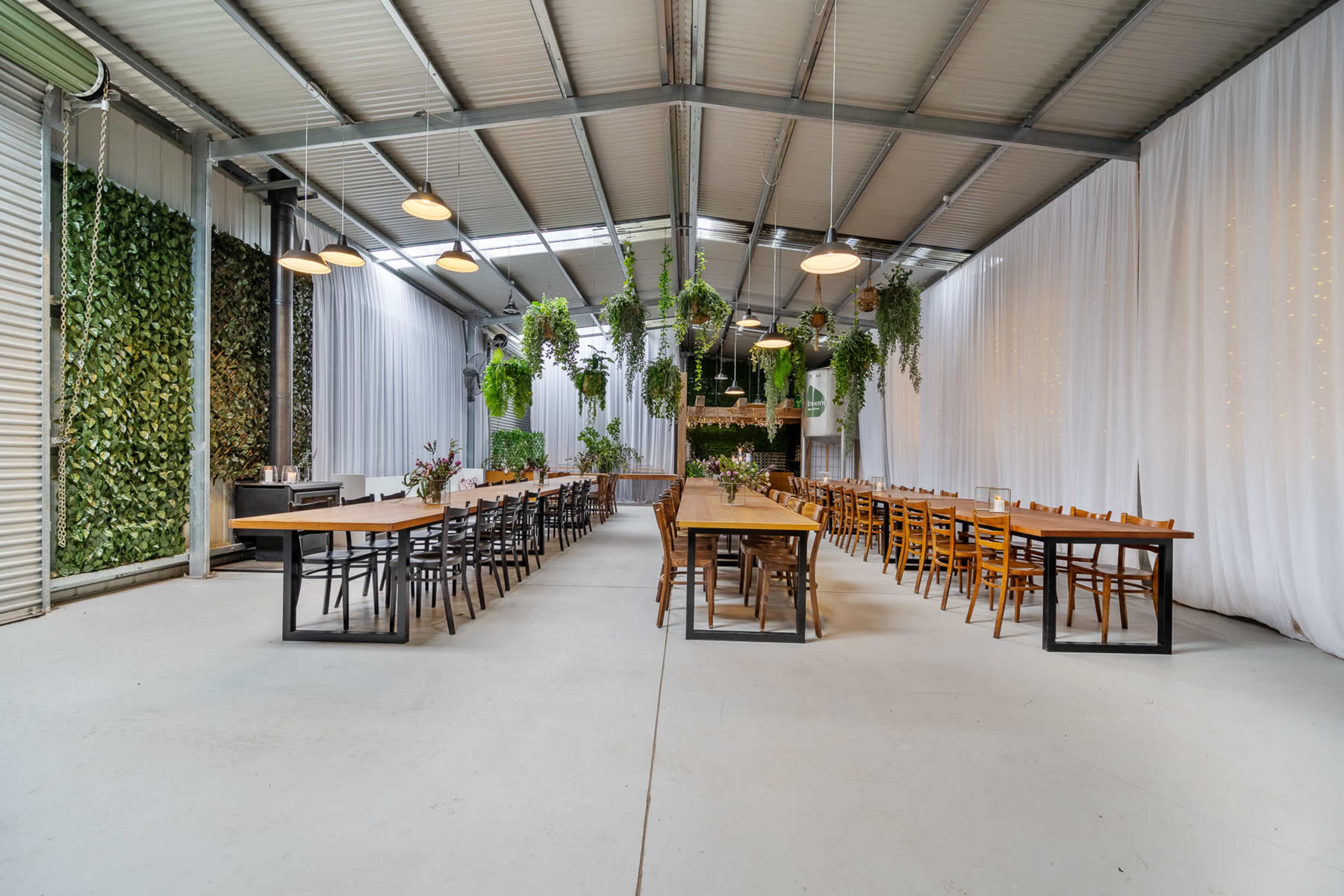 The image shows a spacious indoor venue with long wooden tables arranged in rows, surrounded by white drapery and greenery hanging from the ceiling.
