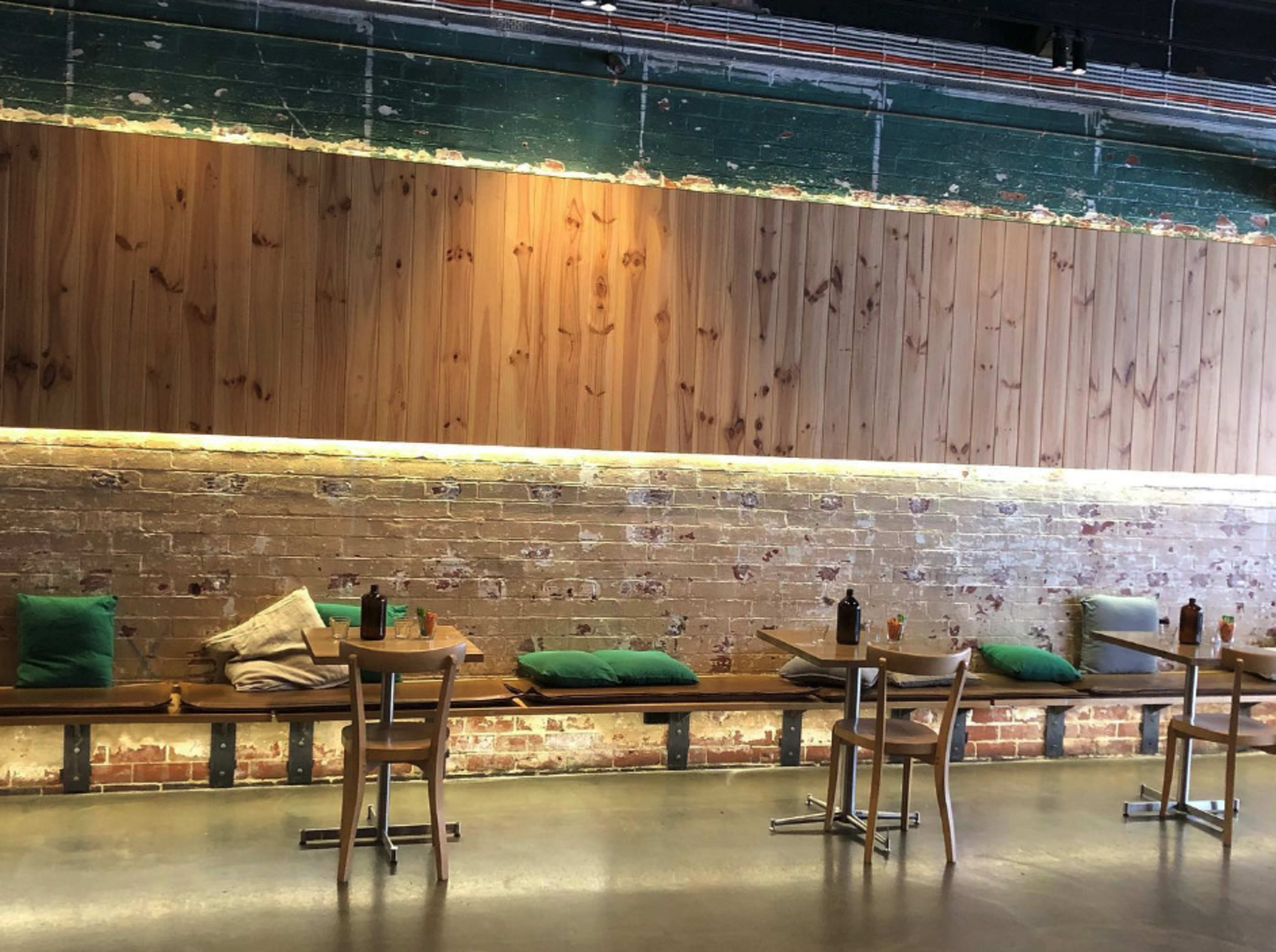 A rustic restaurant interior with wooden walls, a brick backdrop, and wooden tables equipped with green cushions and condiments.