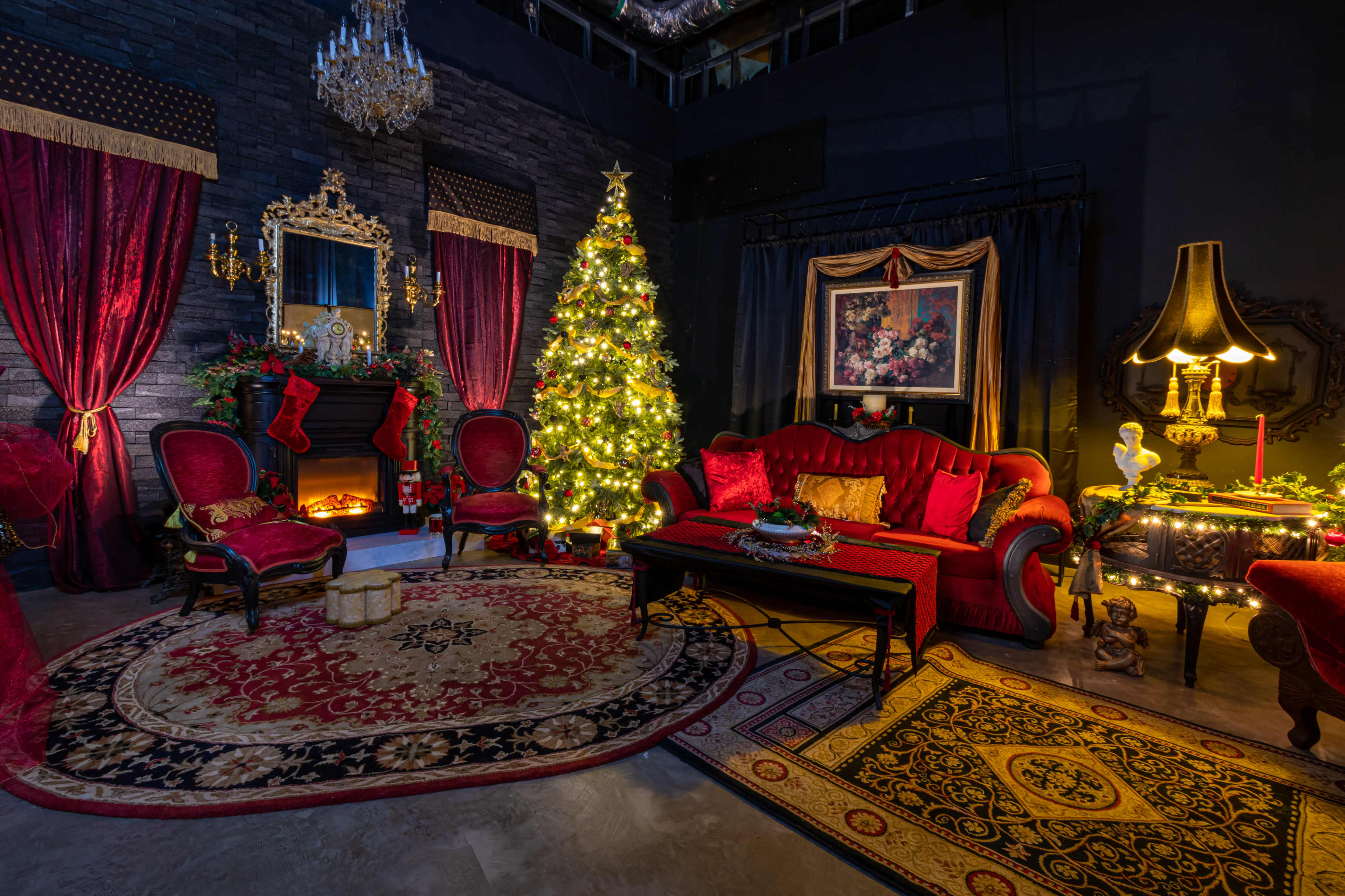 A festive living room decorated with a Christmas tree, a fireplace, and elegant red furniture, all set against dark walls.
