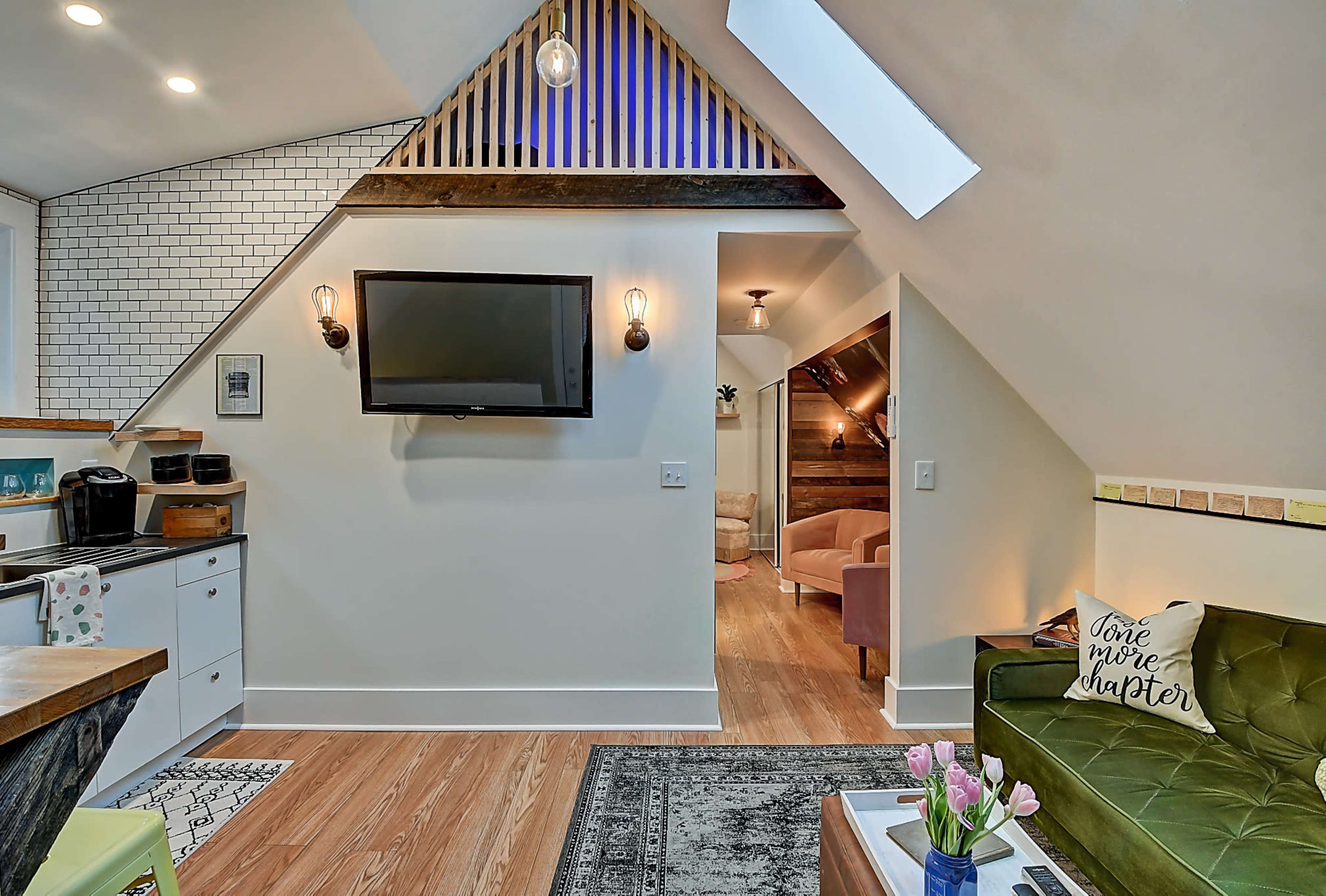 The image shows a cozy living room area with a television mounted on the wall, a green sofa, and a small kitchenette in a bright, well-designed space.