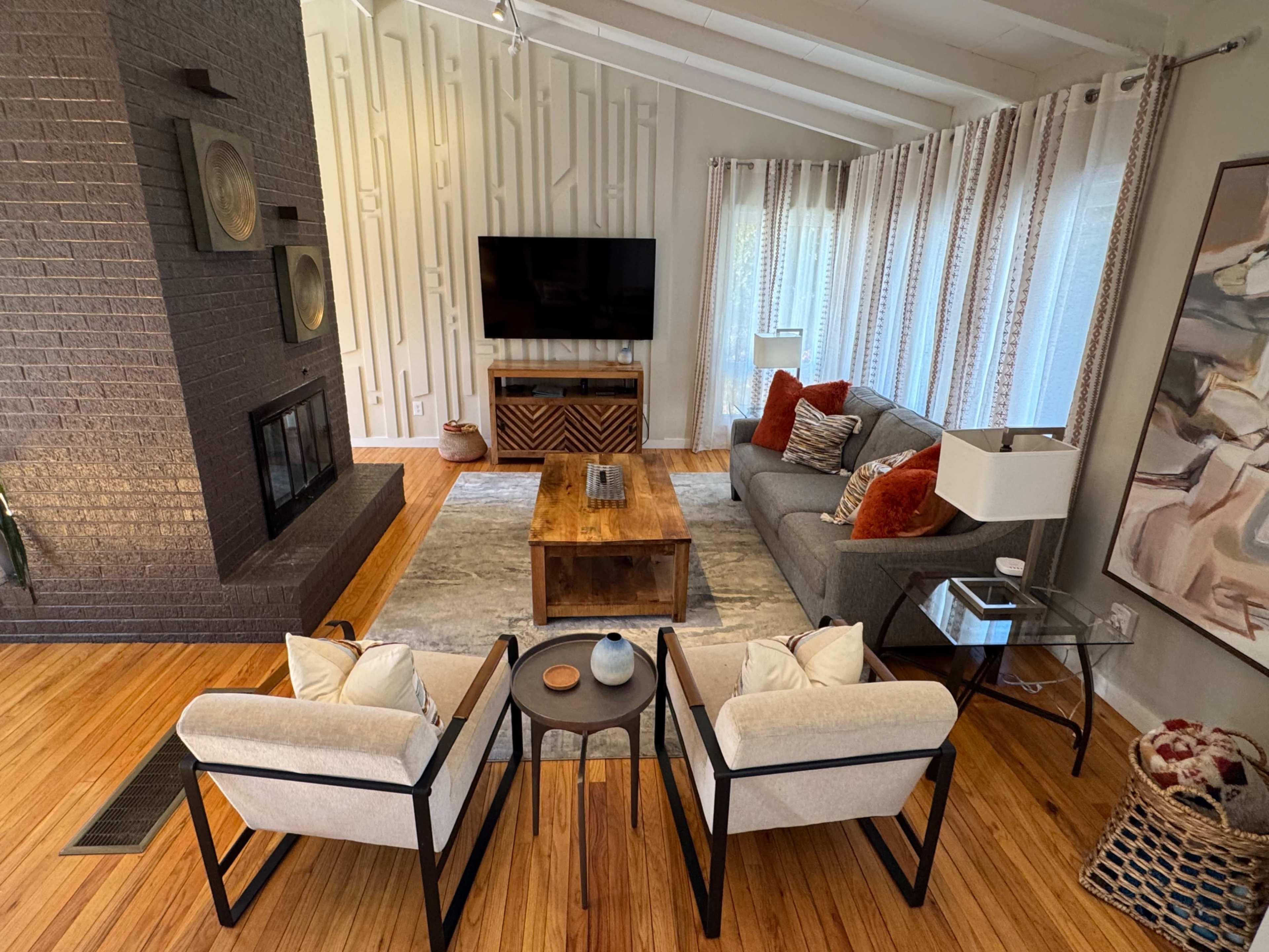 The image shows a modern living room with a gray sofa, two armchairs, a wooden coffee table, a television mounted on the wall, and large windows draped with sheer curtains.