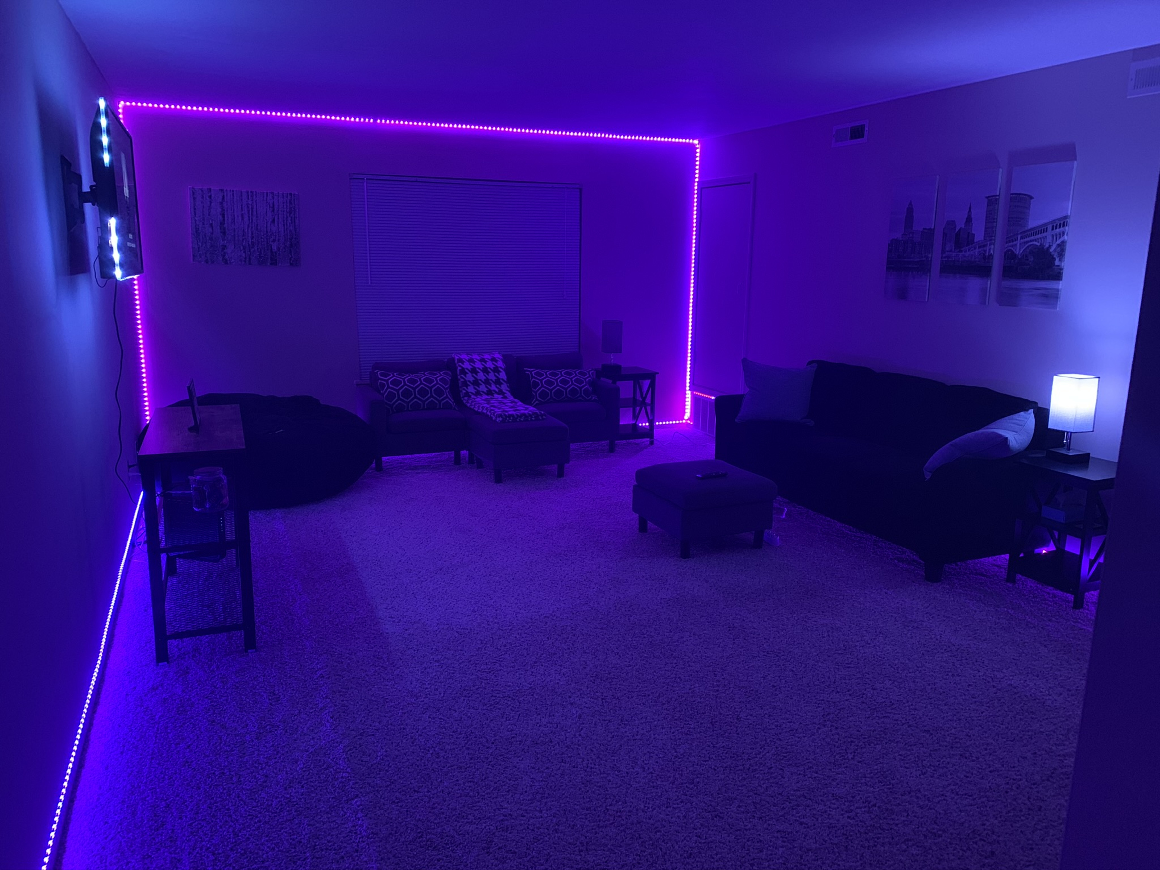 A living room features a purple LED light strip outlining the walls, with a couch, ottoman, and various seating arrangements in a carpeted space.