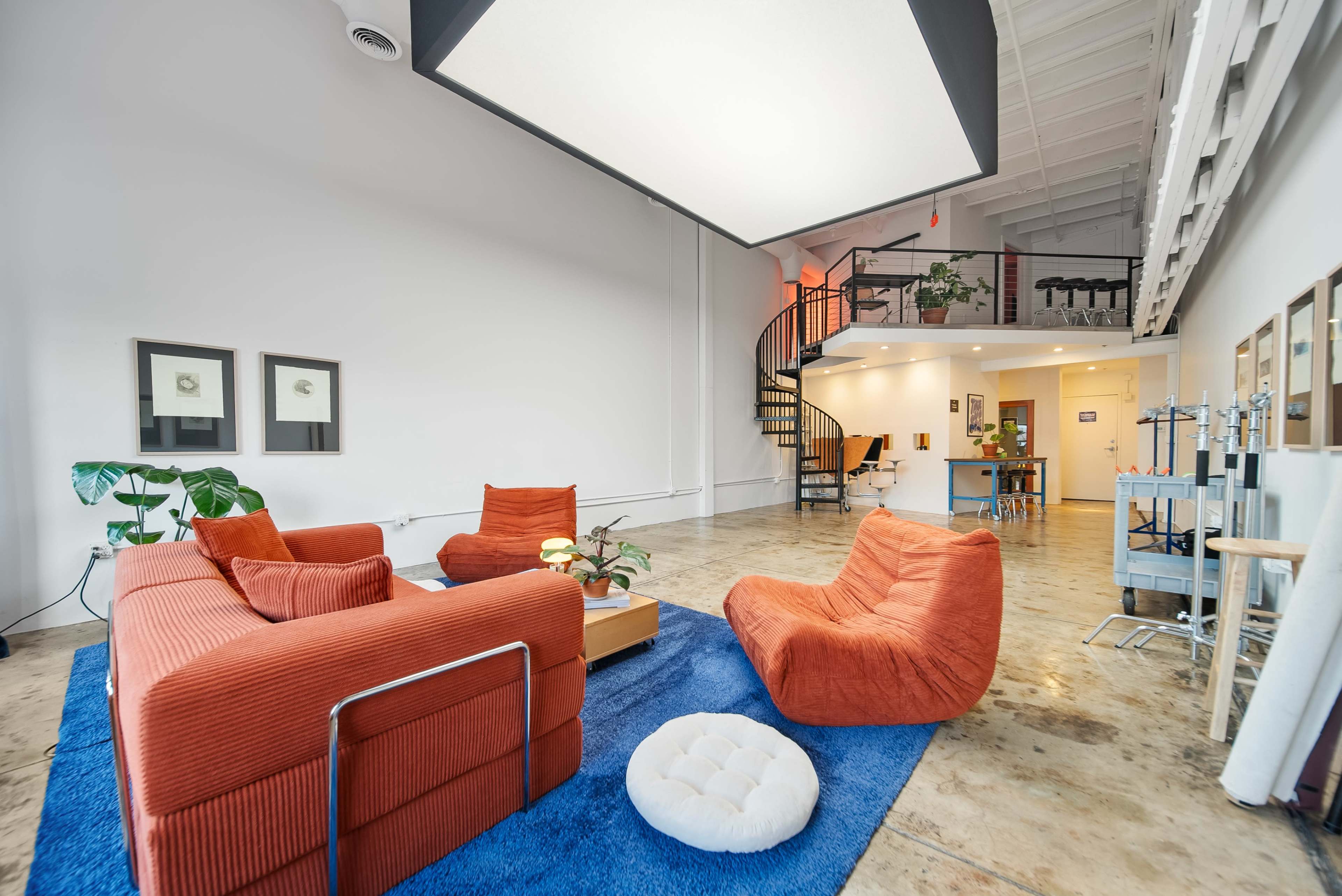 A bright, modern workspace features a cozy seating area with orange sofas, a blue rug, and a spiral staircase leading to an upper level.