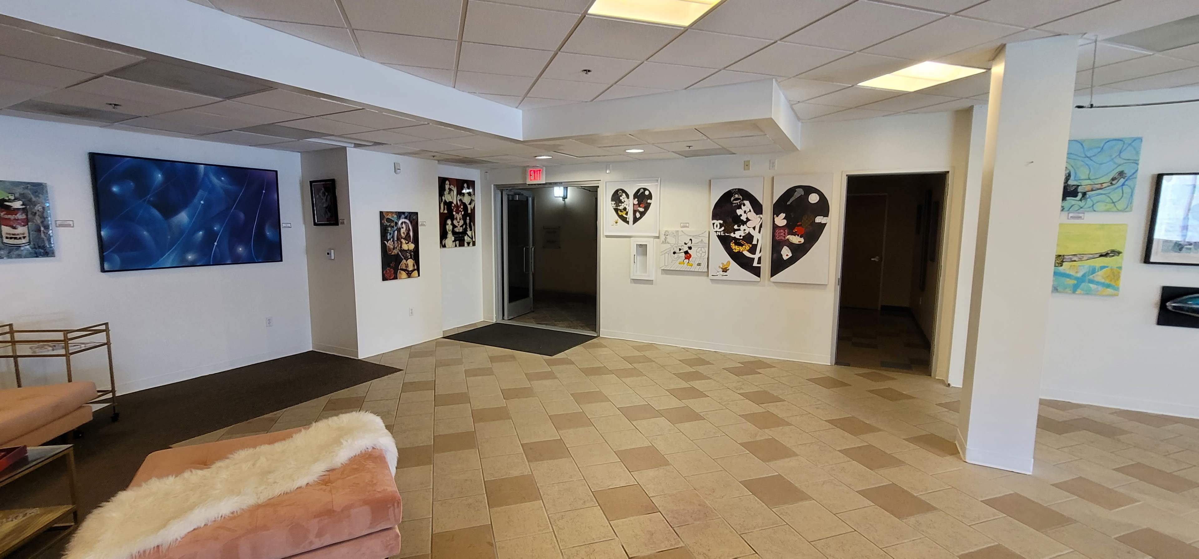 A spacious lobby area with tiled flooring, featuring a reception area, a series of artworks on the walls, and a large television mounted on one wall.