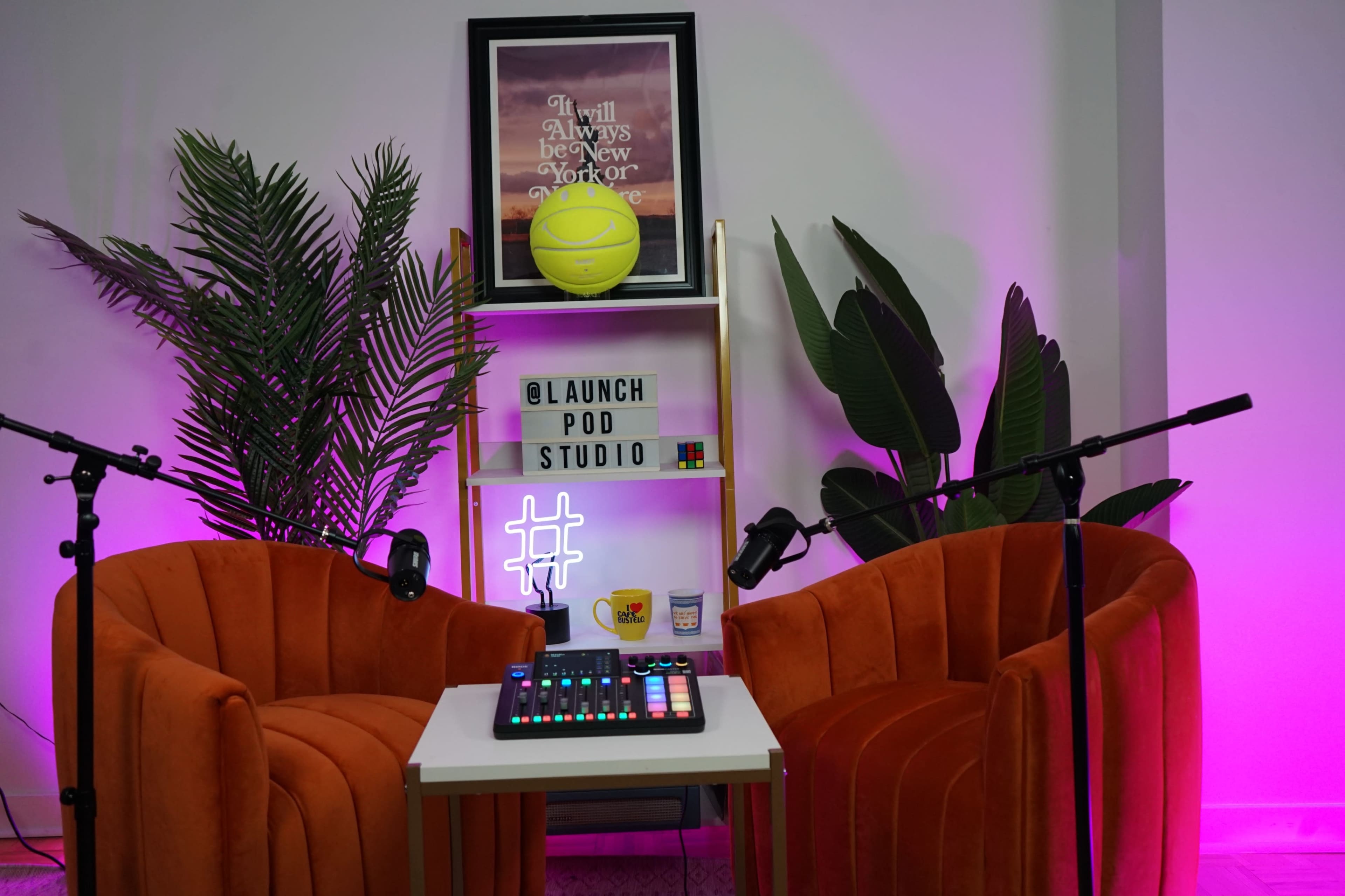 The image shows a podcast studio with two orange velvet chairs, a small table featuring audio equipment, and colorful plants in the background.