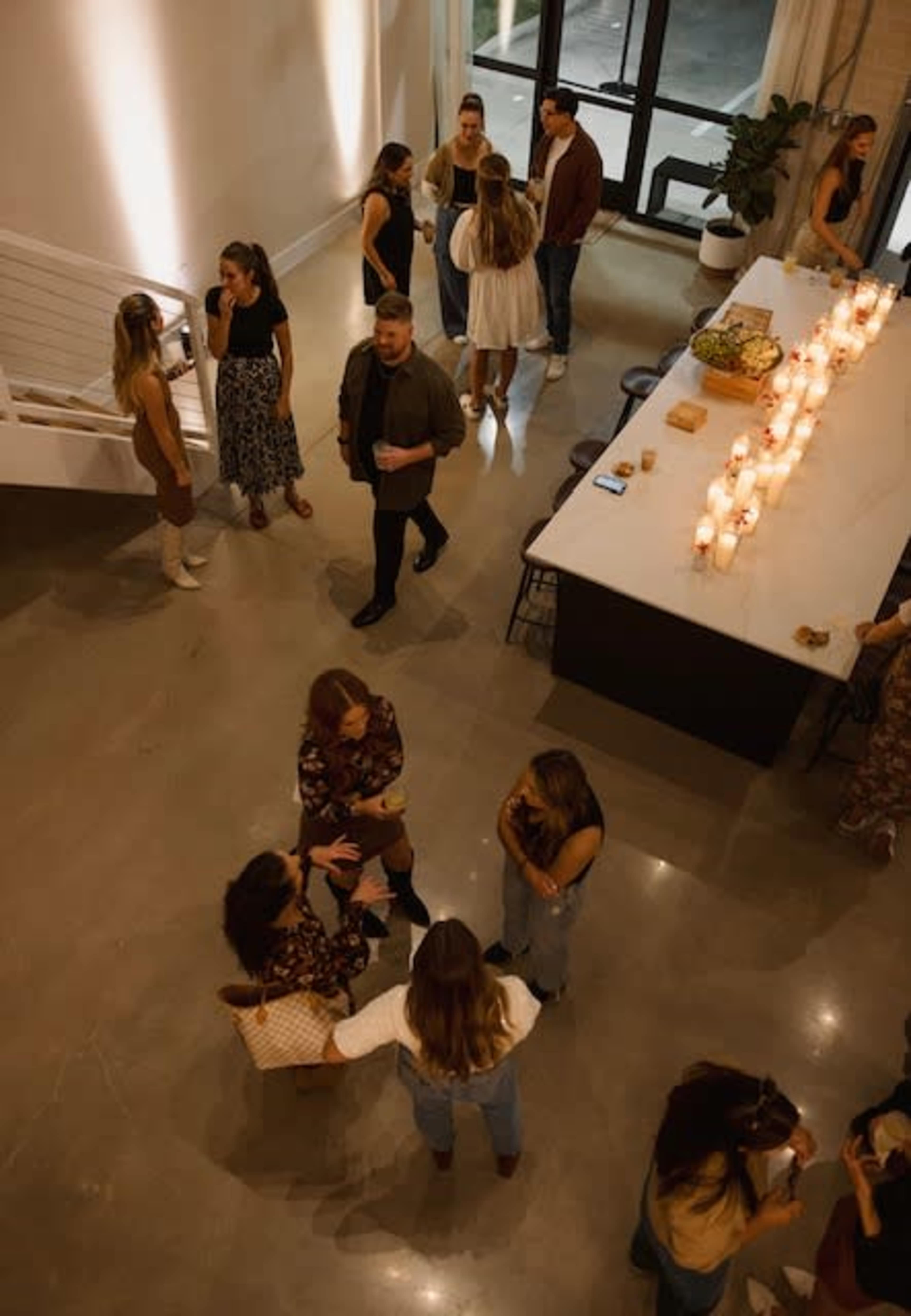 A group of people socializes in a spacious indoor setting with a long table, candles, and a staircase in the background.
