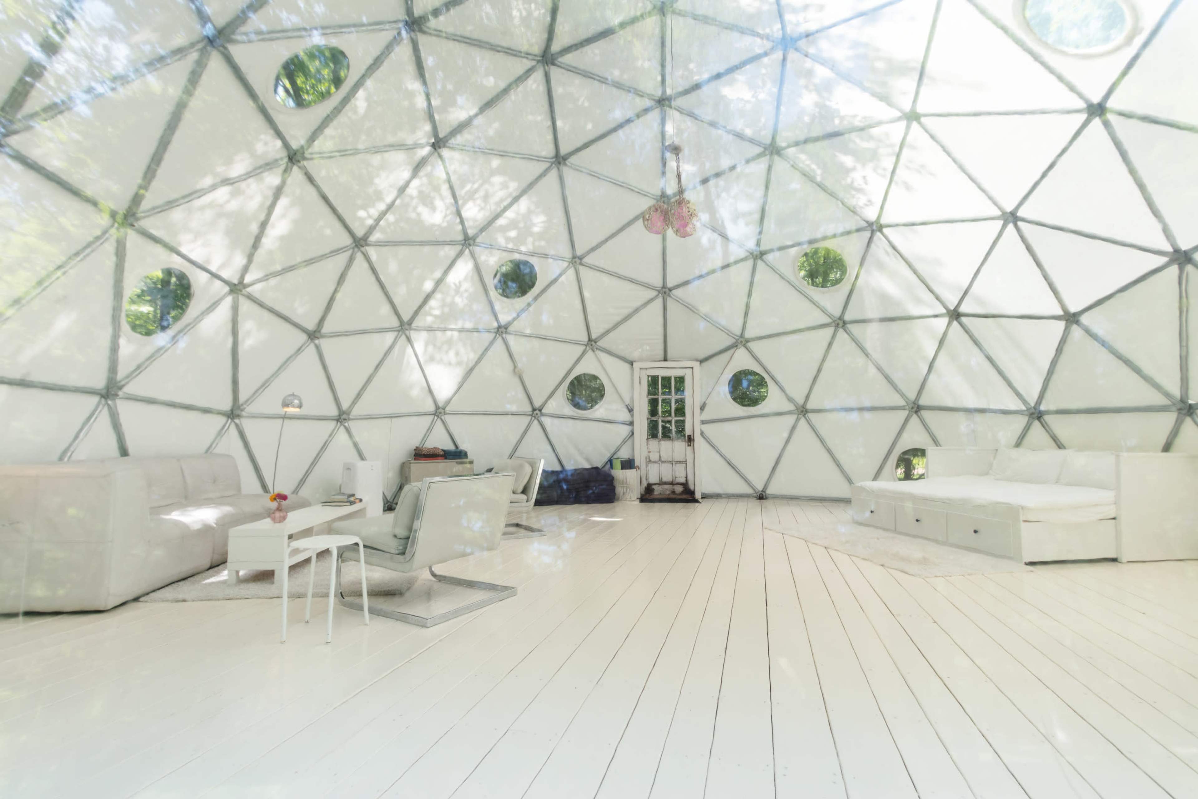 The interior of a geodesic dome features white walls and flooring, with a few minimalistic furniture pieces and large circular windows.